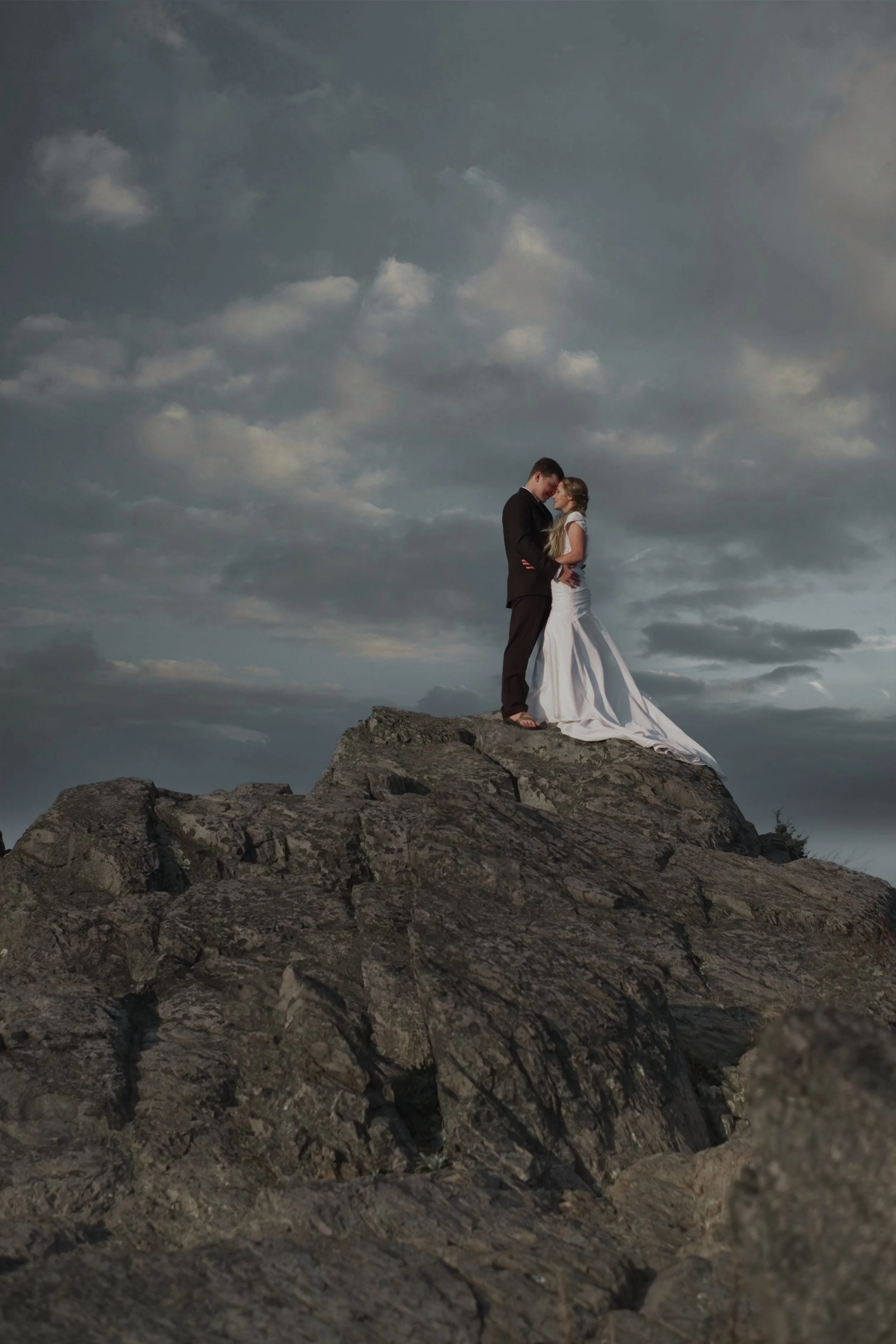 A couple dressed in formal wedding attire standing close together on a rocky hilltop under a cloudy sky with dark, dramatic clouds.