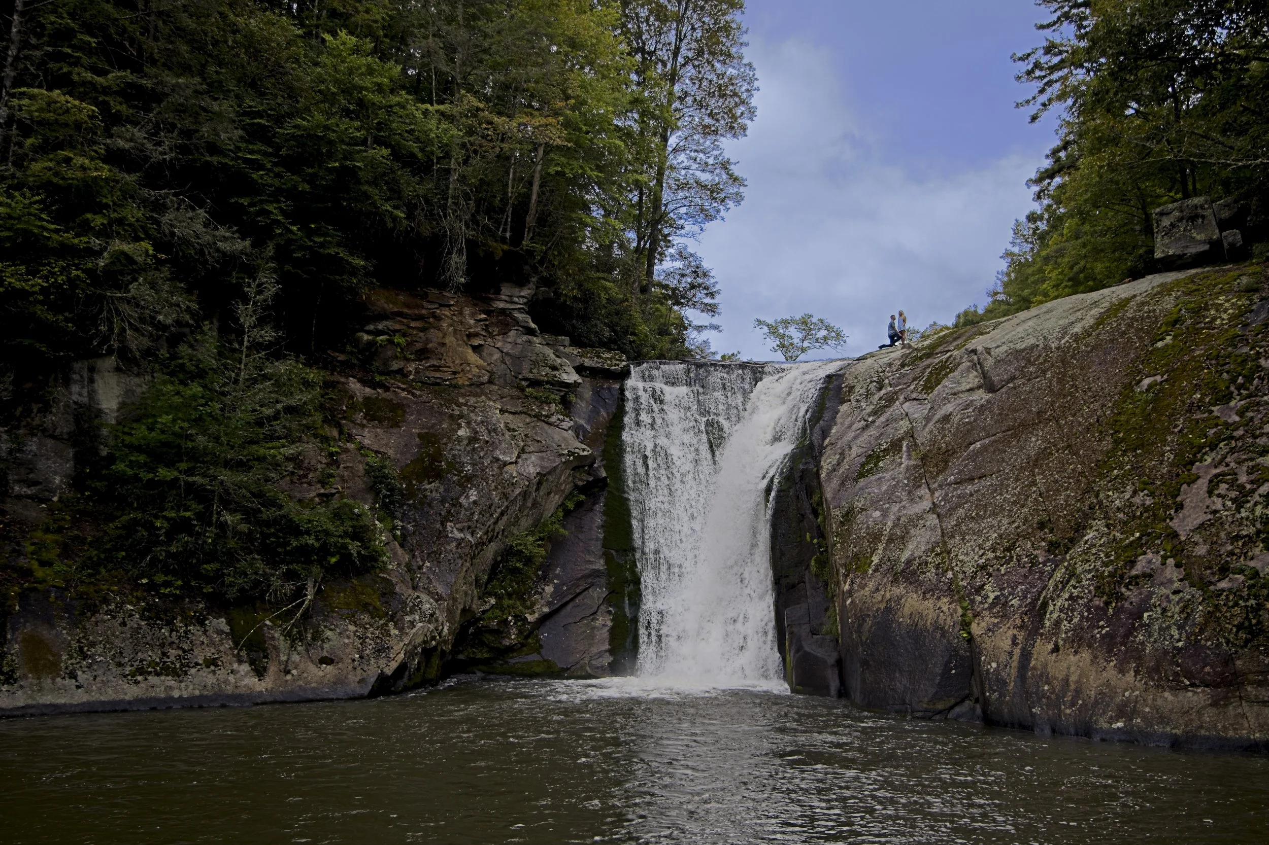 Two people kneeling on rocks near a waterfall in a forested area with a partly cloudy sky.