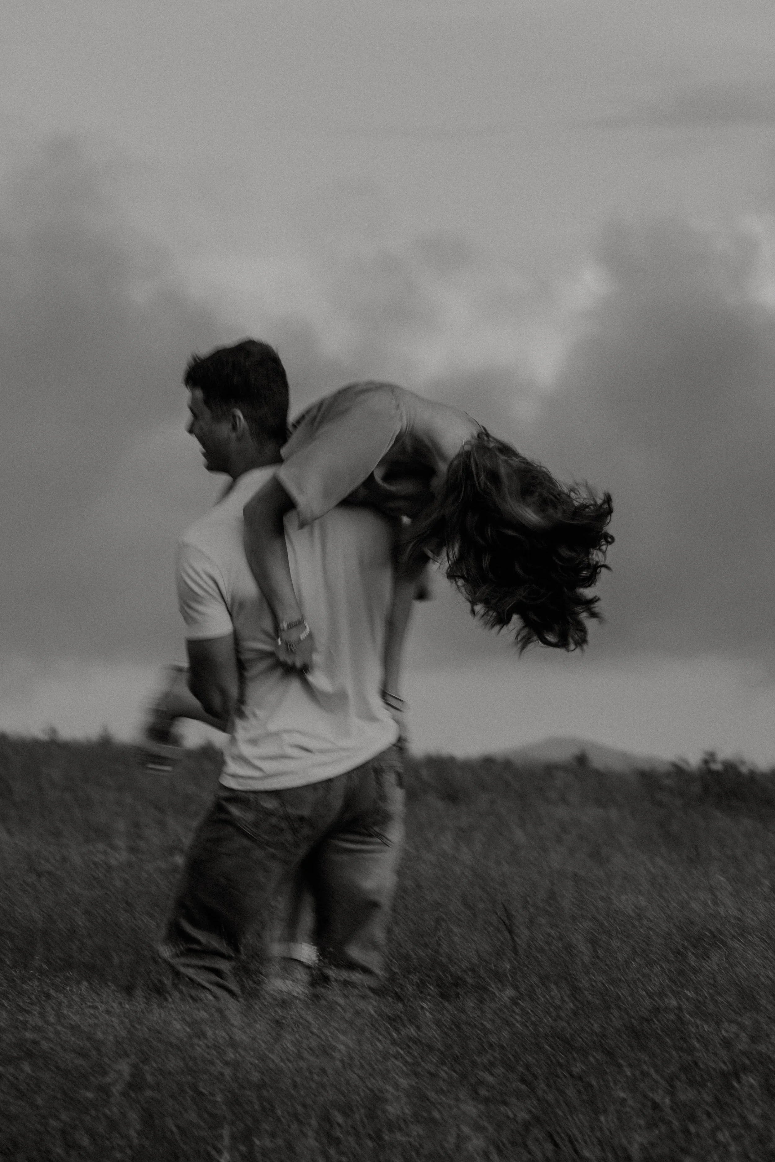 A black and white photo of a man and woman outdoors, with the woman on his back, in a field under cloudy sky.