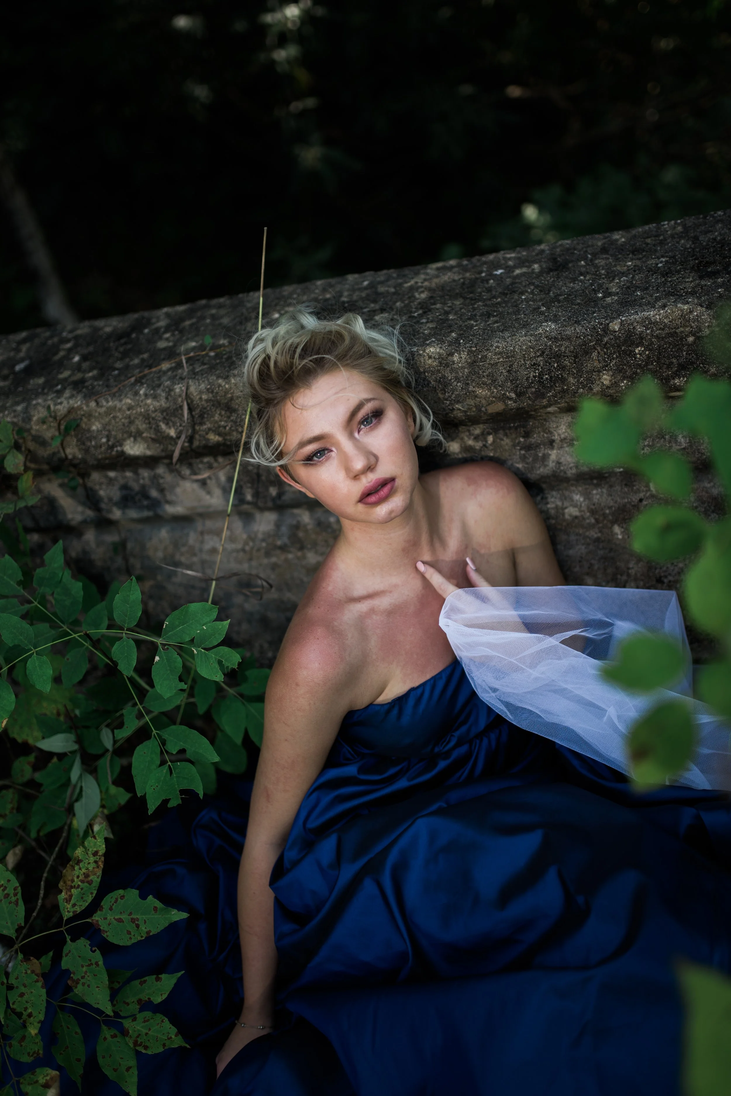 Young woman with platinum blonde hair styled in loose waves, wearing a strapless navy dress with a tulle accent, lying against a stone wall outdoors amidst green foliage, gazing into the camera with a neutral expression.