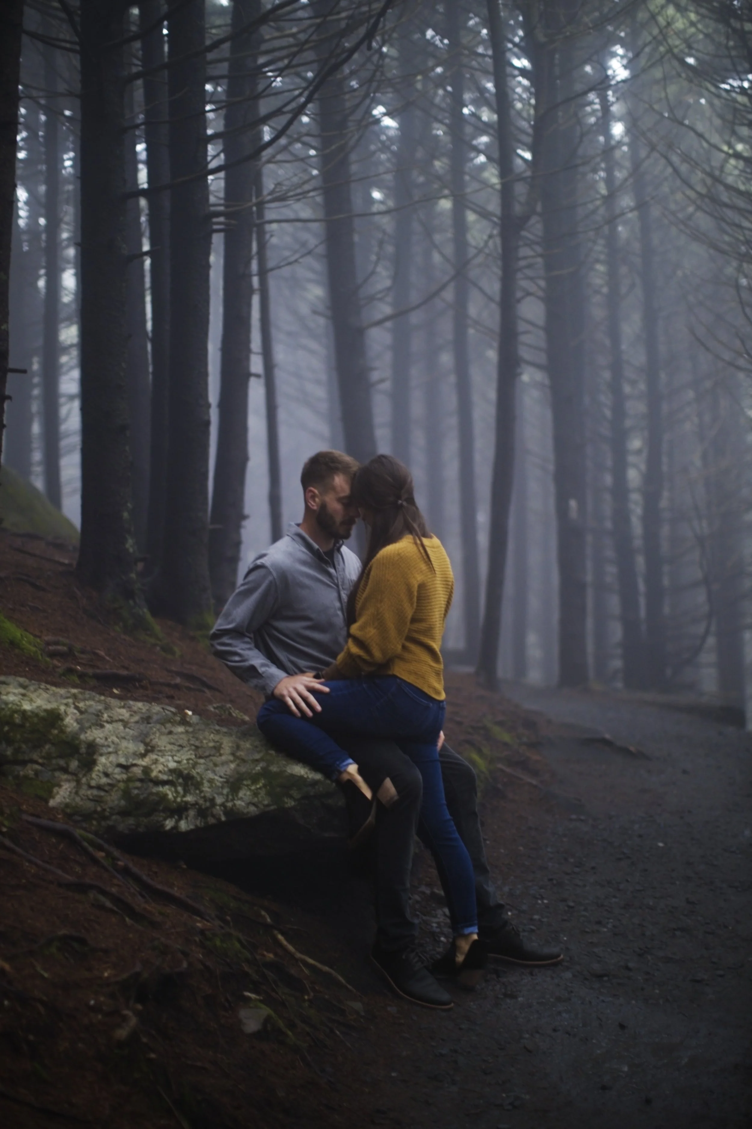 A man and woman sitting close together on a mossy log in a misty forest, embodying a romantic moment.