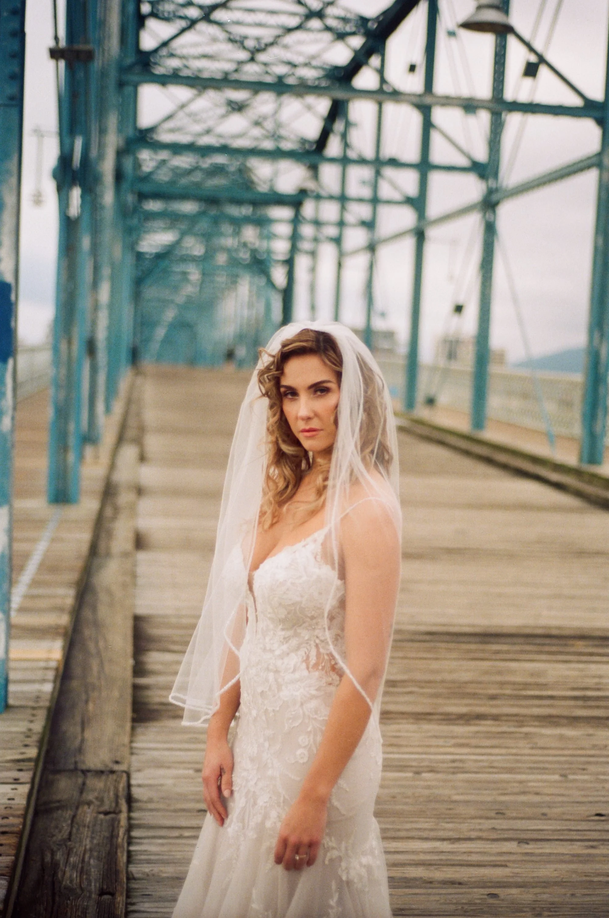 A woman in a white wedding dress with lace details and a veil standing on a wooden bridge with blue metal framing and background of cloudy sky.