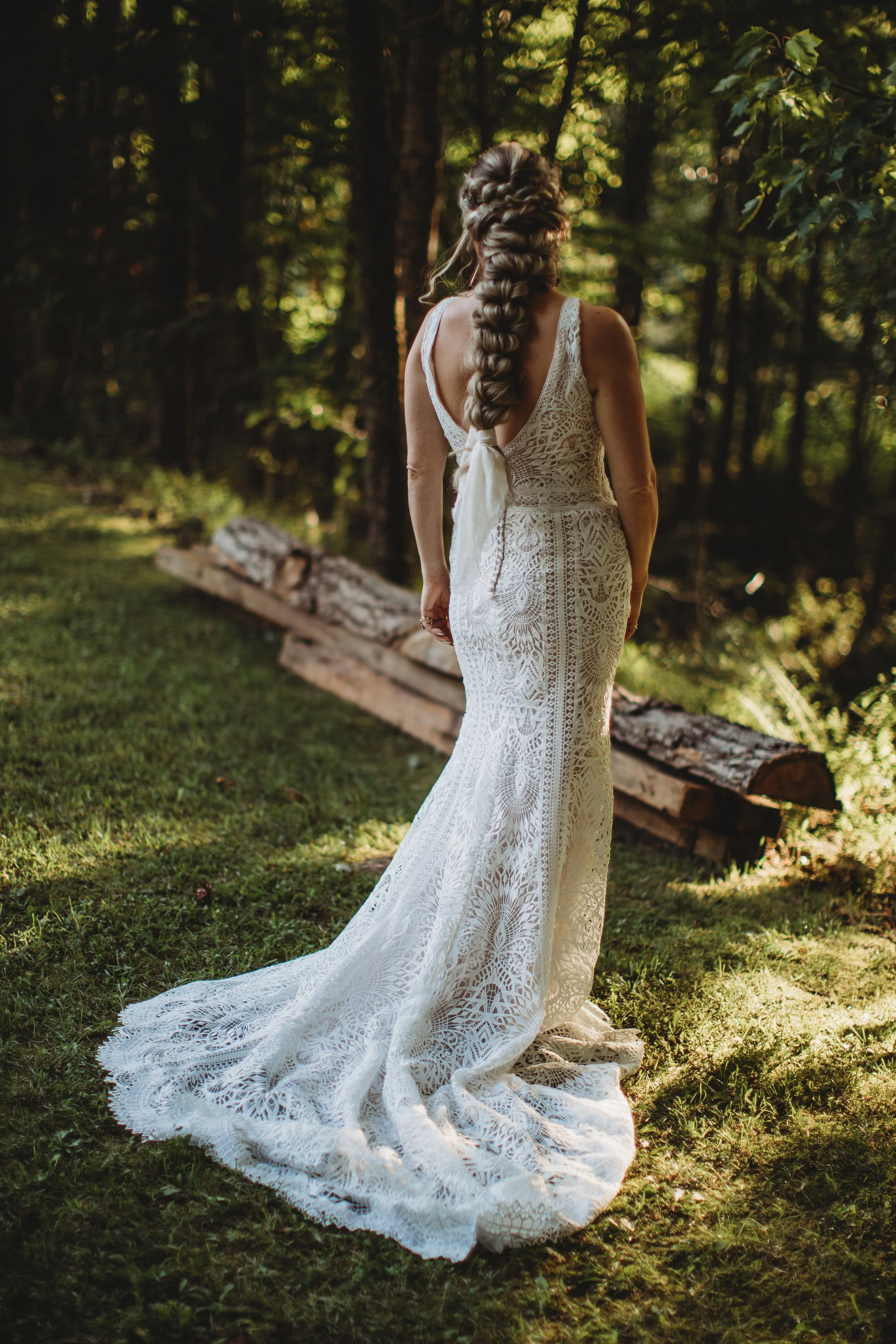 A woman wearing a long, lace white dress with a train stands outdoors in a wooded area, facing away from the camera, with her hair styled in a large braid.