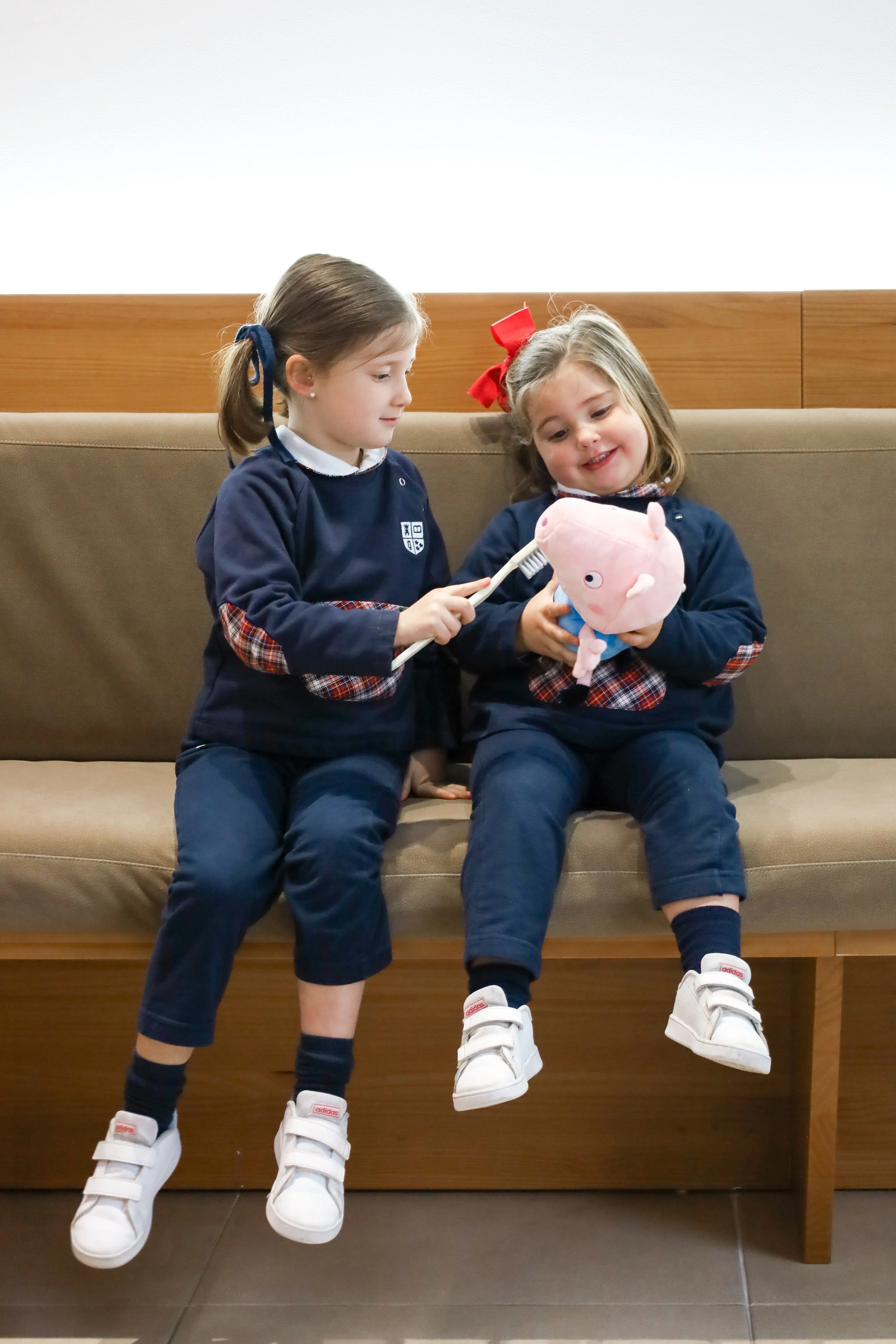 Two girls in school uniform sitting on a sofa, one of them holding a stuffed toy while the other appears to brush it with a toothbrush.