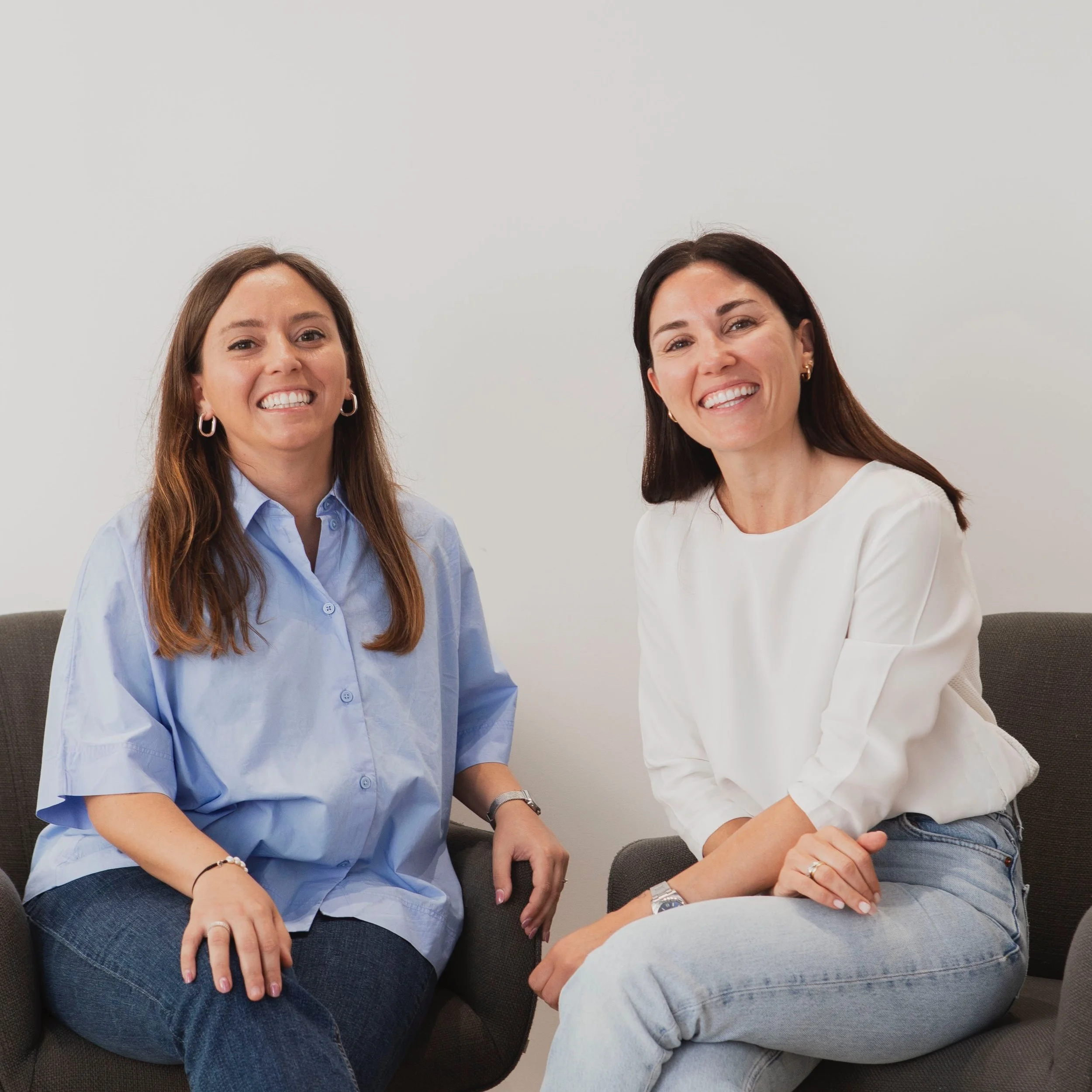 Dos mujeres sentadas en un sillón, sonríen y parecen estar conversando en un ambiente de oficina.