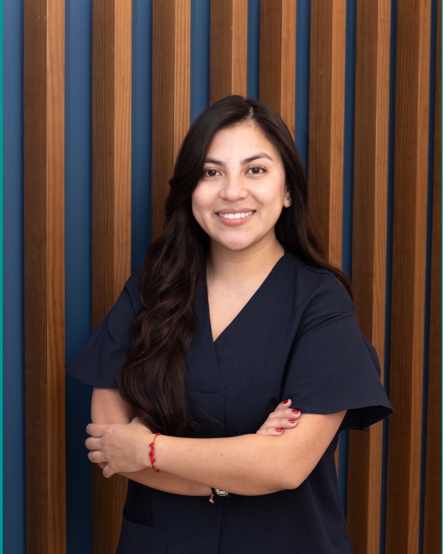 Mujer de cabello largo y oscuro, con uniforme azul, sonriendo frente a un fondo de paneles de madera y azul.