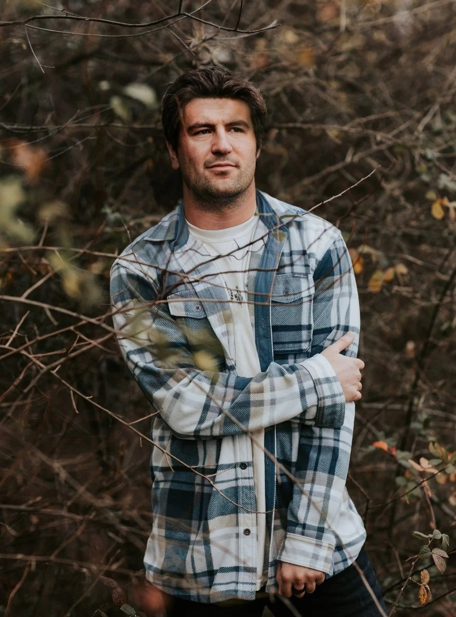 A young man with dark hair and a five o'clock shadow standing outdoors among leafless branches, wearing a plaid shirt over a white t-shirt.