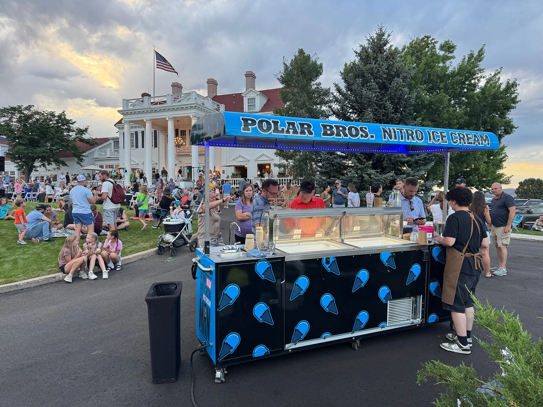 A Polar Bros. Nitro Ice Cream cart with a blue canopy, selling ice cream at an outdoor event with many people, a white house, trees, and an American flag in the background.