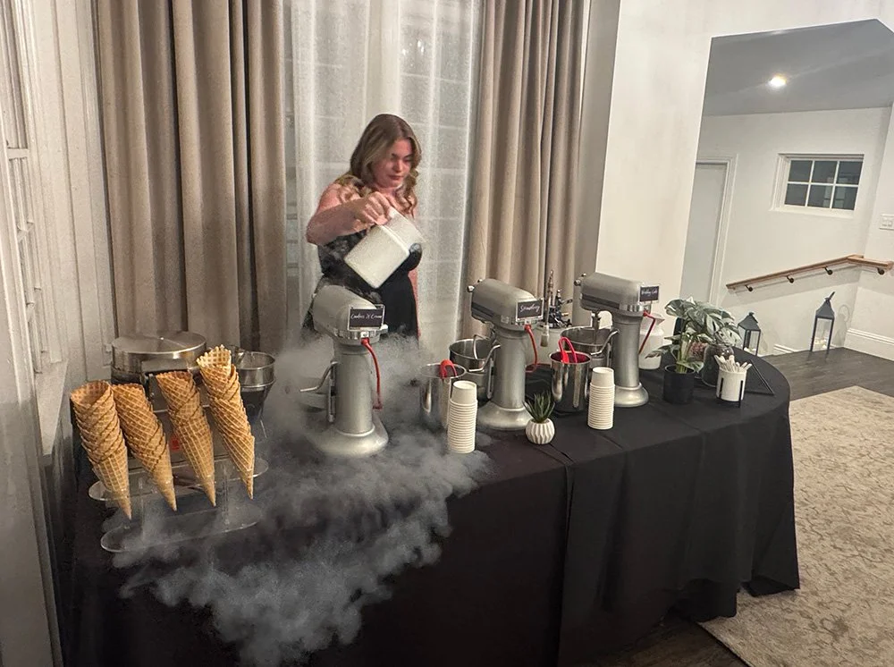 A woman working behind a Polar Bros Nitro Ice Cream table at a wedding event with three ice cream makers, cones, and cups, using liquid nitrogen and creating vapor.