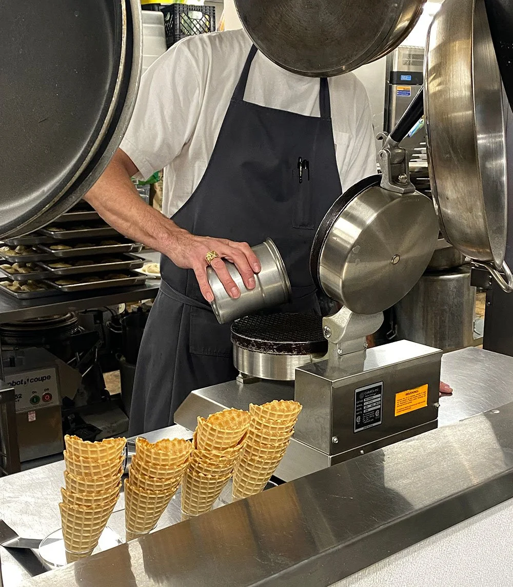 Polar Bros Nitro Ice Cream team member wearing a white shirt and black apron making waffle cones with a waffle cone iron. Four waffle cones are stacked on a tray in the foreground.