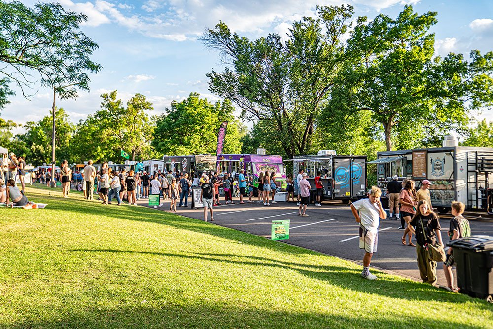 Line of food trucks at an outdoor event with a large crowd of people, green trees, and a grassy area under a blue sky.