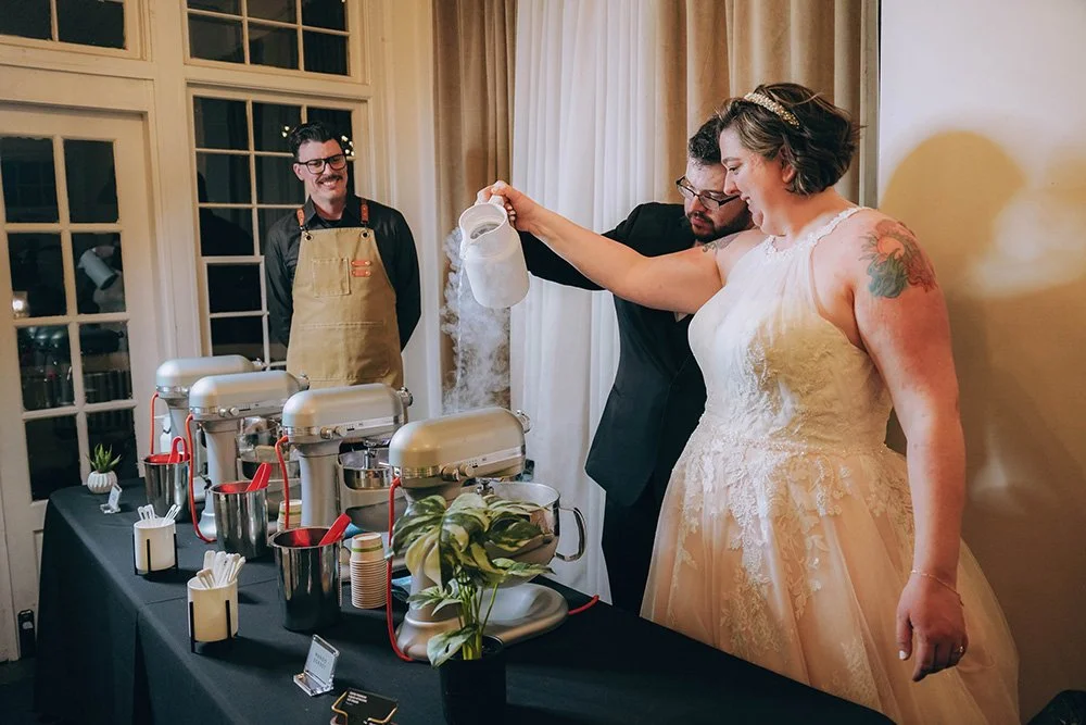 A bride with a tattoo on her arm wearing a wedding dress pours liquid nitrogen into a bowl, overseen by Chef Chuck James, Owner of Polar Bros Nitro Ice Cream during a wedding celebration.