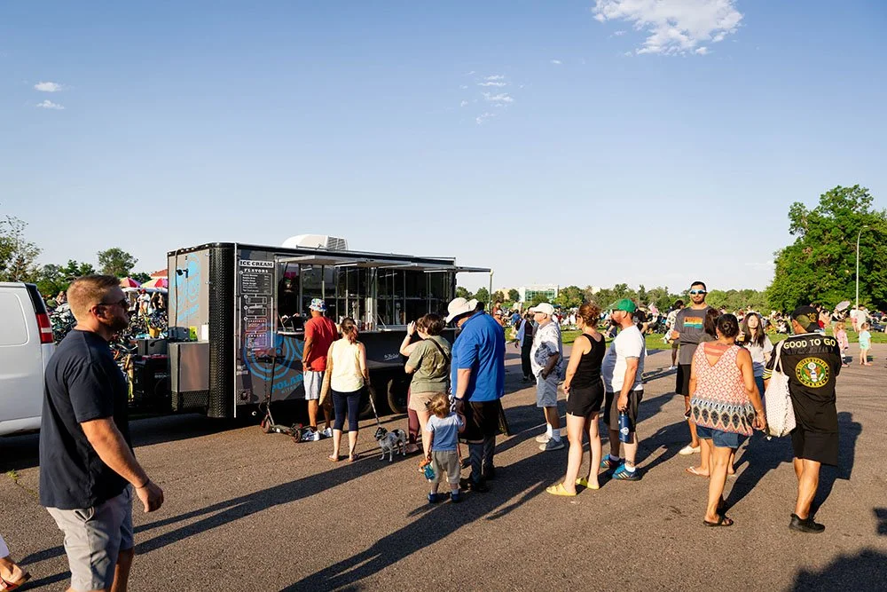 People line up at a Polar Bros Nitro Ice Cream food truck during daytime, with a clear blue sky and trees in the background.