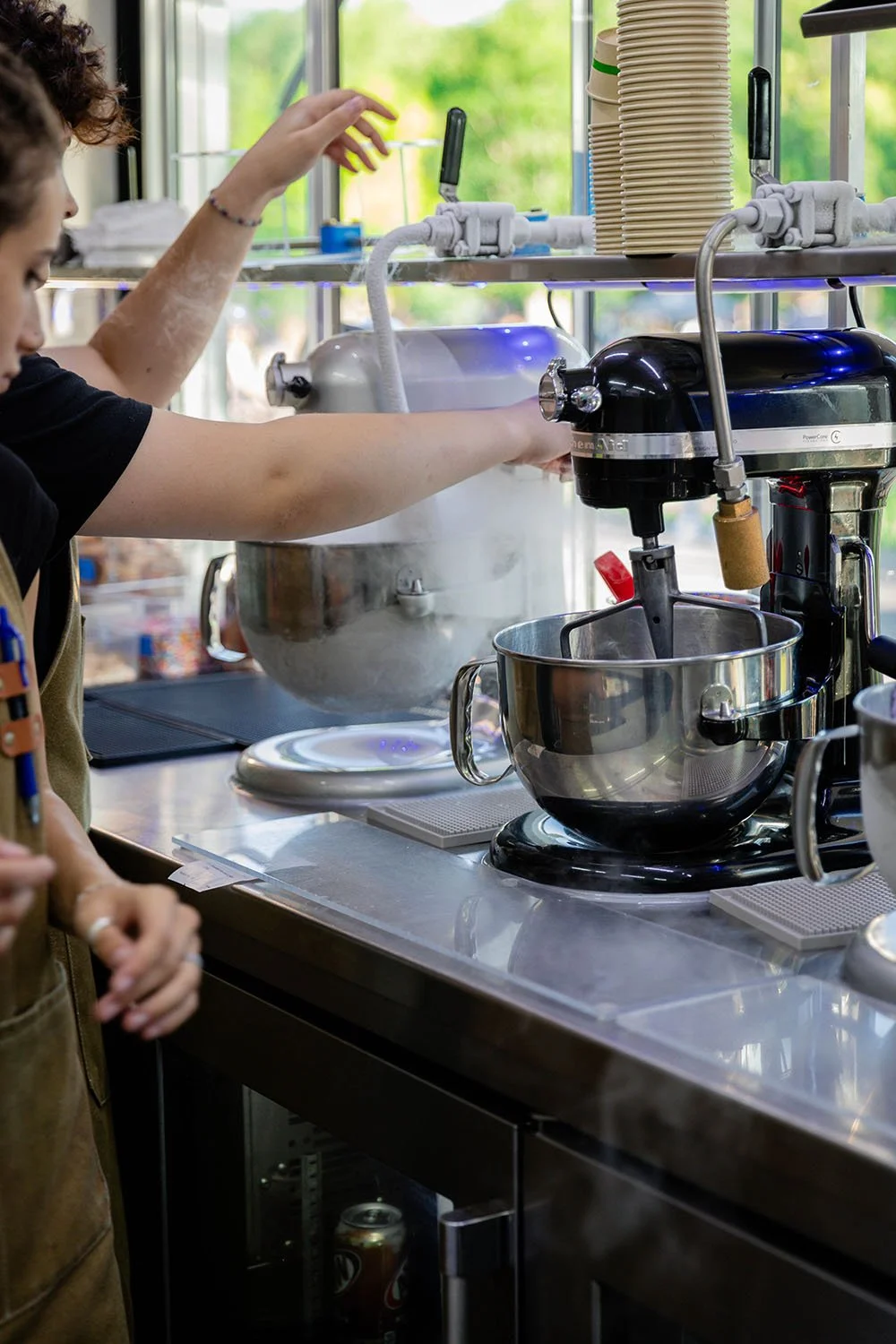 A person operating a commercial stand mixer in a Polar Bros Nitro Ice Cream food truck with a coffee machine nearby and stacks of disposable cups in the background.