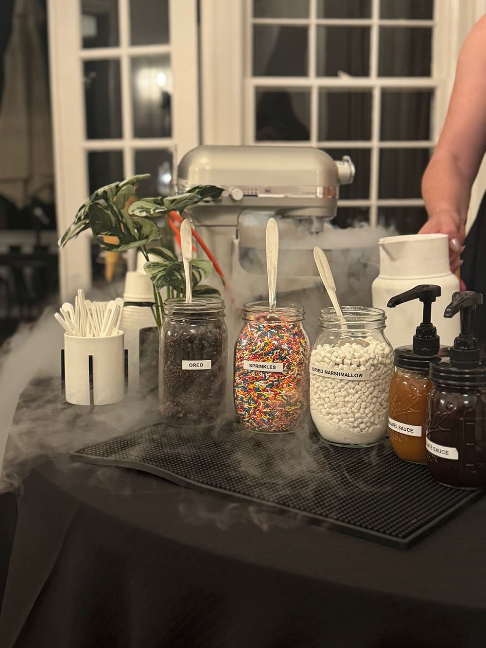 Counter with jars of Oreo cookies, sprinkles, dried marshmallow, caramel sauce, and chocolate sauce, with a mixer in the background and steam rising from the jars.