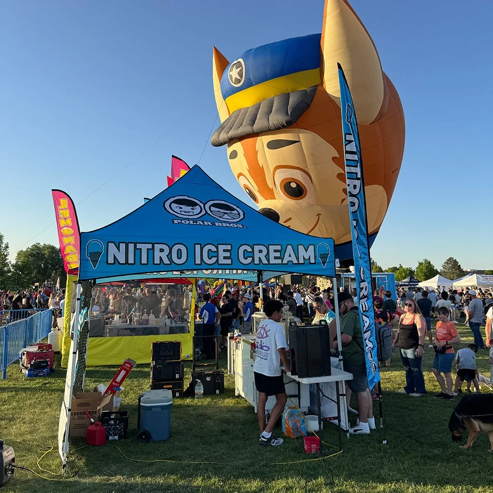 A carnival or fair with a large hot air balloon shaped like a cartoon dog wearing a military hat. In the foreground, there is a booth selling nitro ice cream with a blue canopy and pink flags. There are people gathered around the booth and others wal