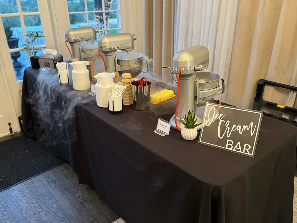Ice cream bar setup with three machines, white containers, cups, spoons, and a sign that says 'Ice Cream Bar'. Vapor from liquid nitrogen is coming from the machines.