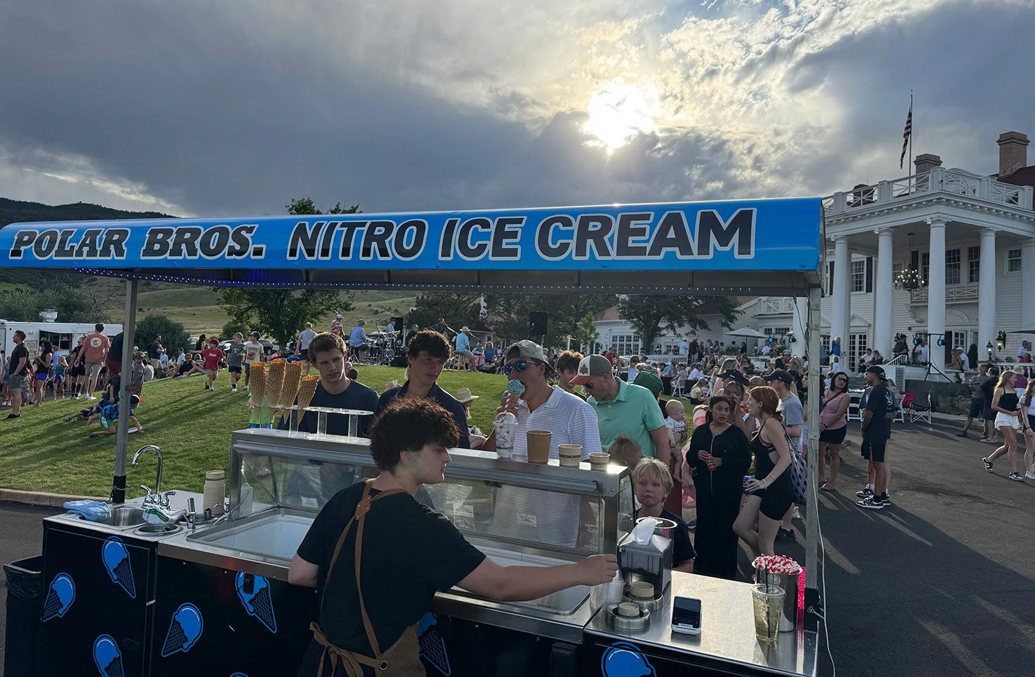 People standing in line at a Polar Bros. Nitro Ice Cream stand during a sunny outdoor event, with a large white building in the background and a cloudy sky overhead.