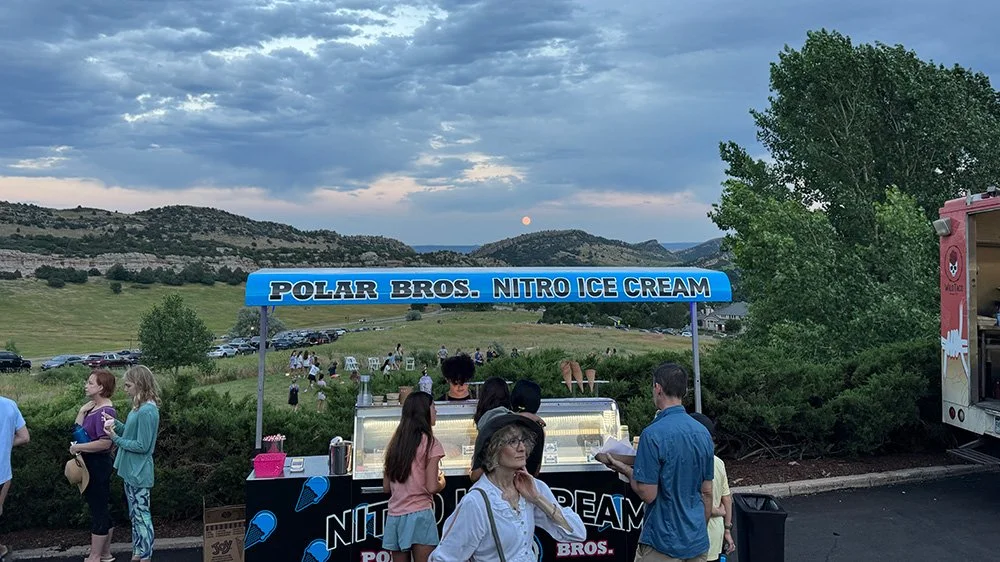 People standing in line at a Polar Bros Nitro Ice Cream cart in an outdoor park setting during evening, with hills and a cloudy sky in the background.