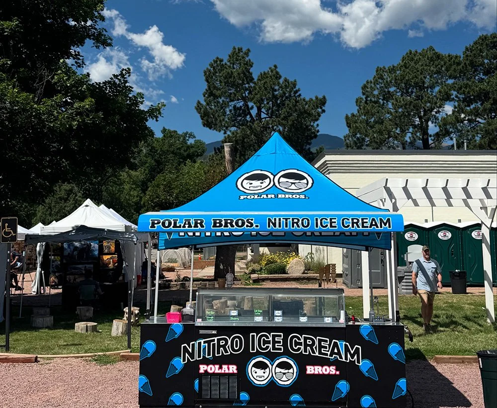 Outdoor ice cream stand with a blue tent labeled 'POLAR BROS. NITRO ICE CREAM' and a black counter with a display case, surrounded by trees and people at an outdoor event.