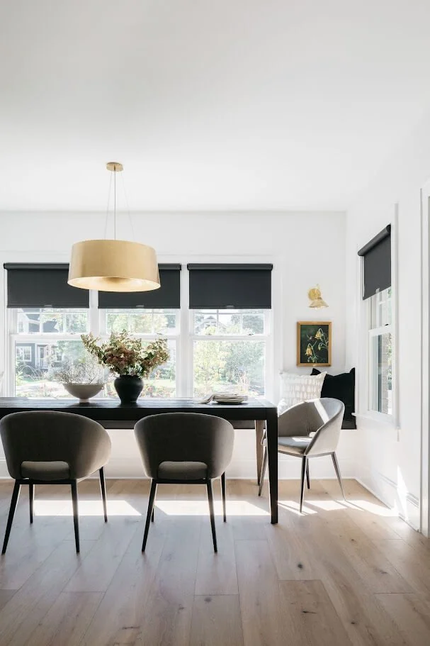 A single neutral roman shade that ofers exceptional light control accents a modern bathroom with black iron shower surround. Bathroom vanity with beautiful accesssories.  The bathroom space is filled with natural light.