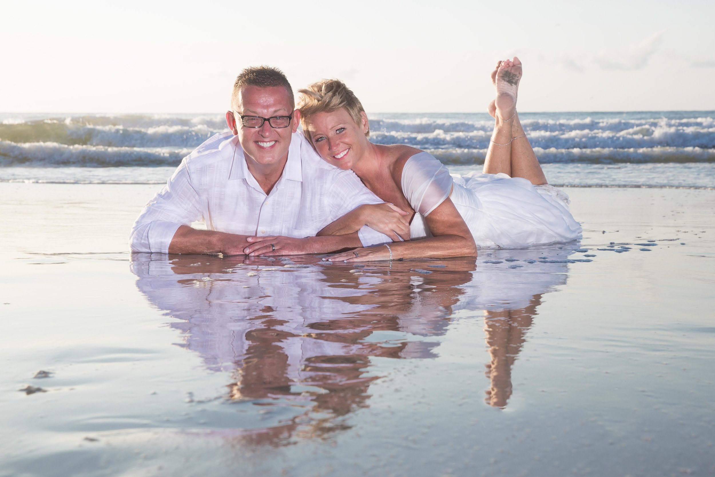  Husband and wife lay in shallow water of the beach smiling together at the camera 