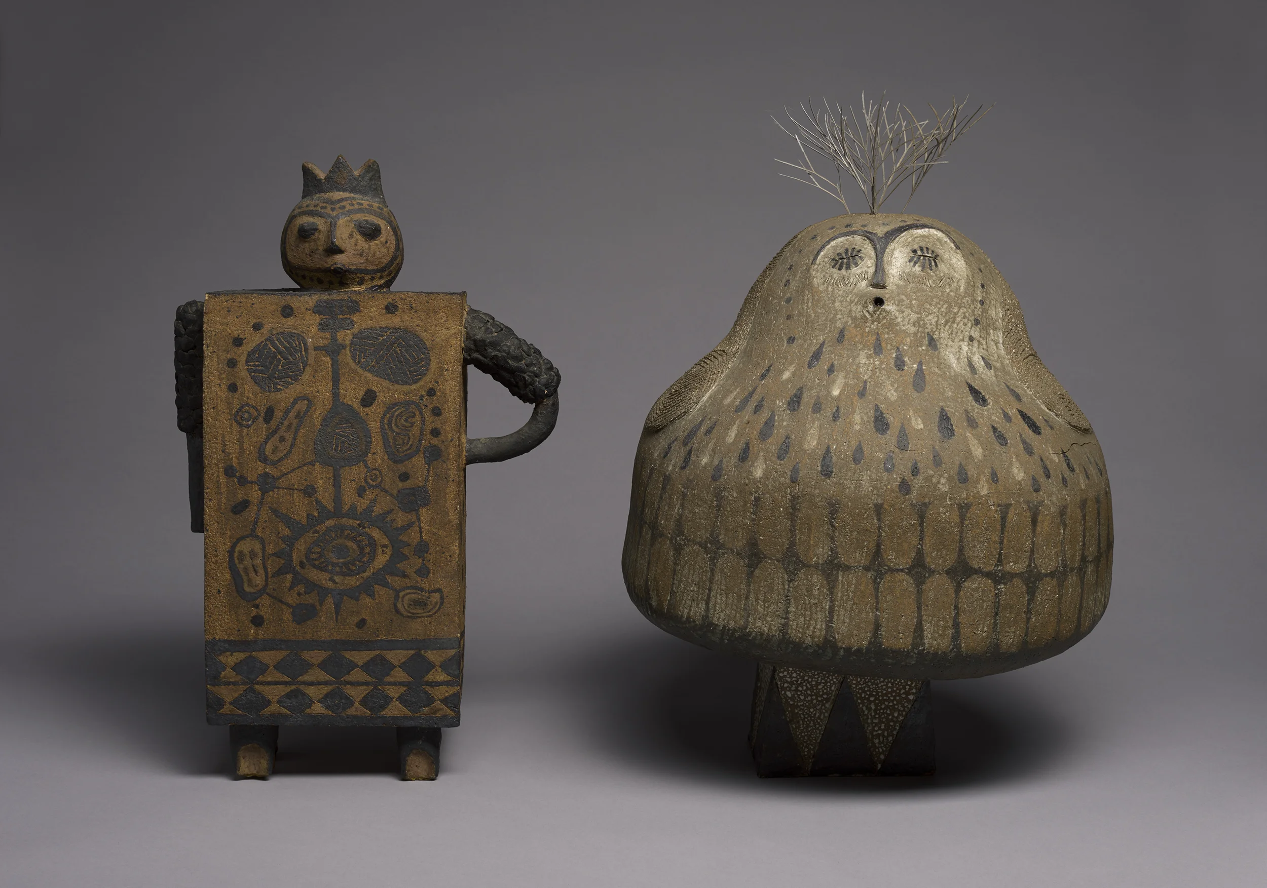 Two traditional African or indigenous art sculptures. The left sculpture is a rectangular figure with a round face, crowned, with detailed patterns on the body, and on the right, a circular, owl-like figure with closed eyes, a beak, and a plant or br