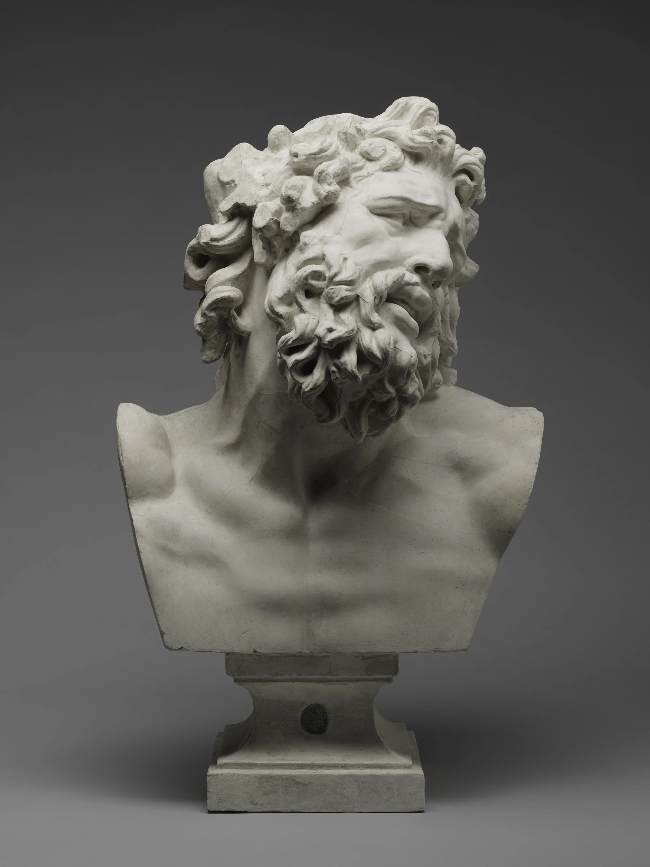 Classical marble sculpture of a male head with a beard and curly hair, displayed on a pedestal against a gray background.