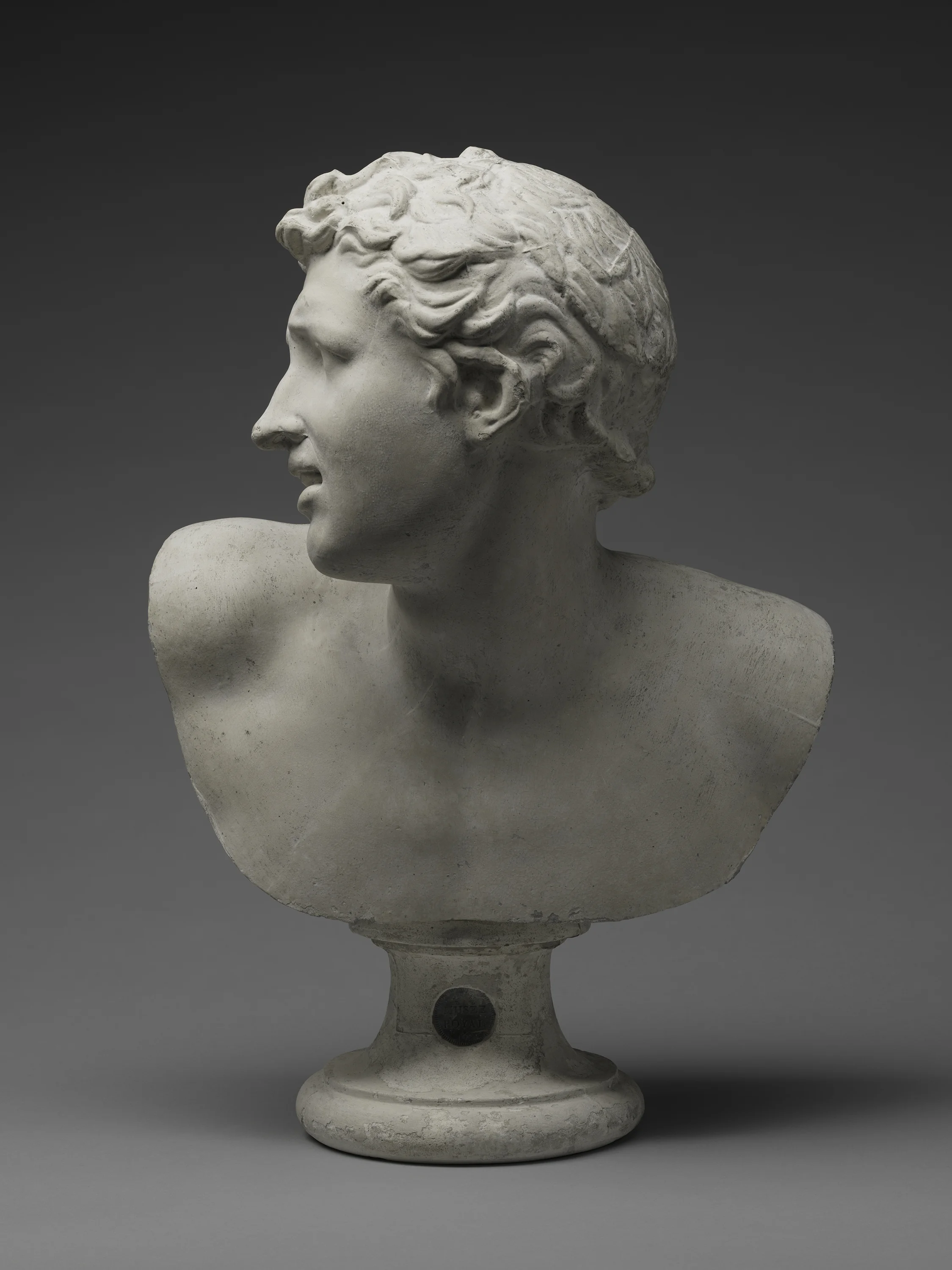 A white marble bust sculpture of a woman with detailed hair, mounted on a pedestal with a circular plaque, against a gray background.