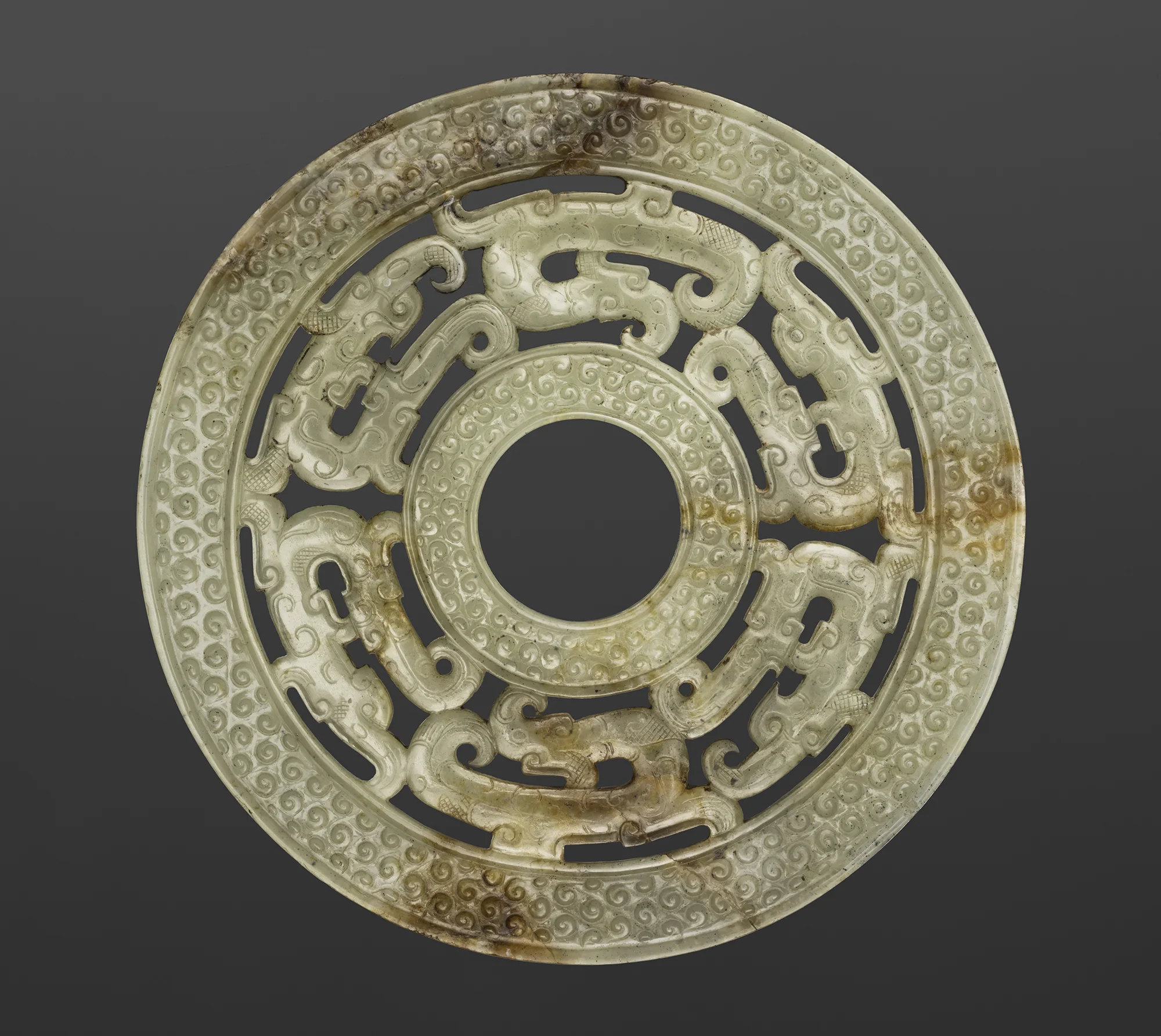A carved circular jade ornament with intricate swirling and mythical creature patterns, aged and slightly discolored.