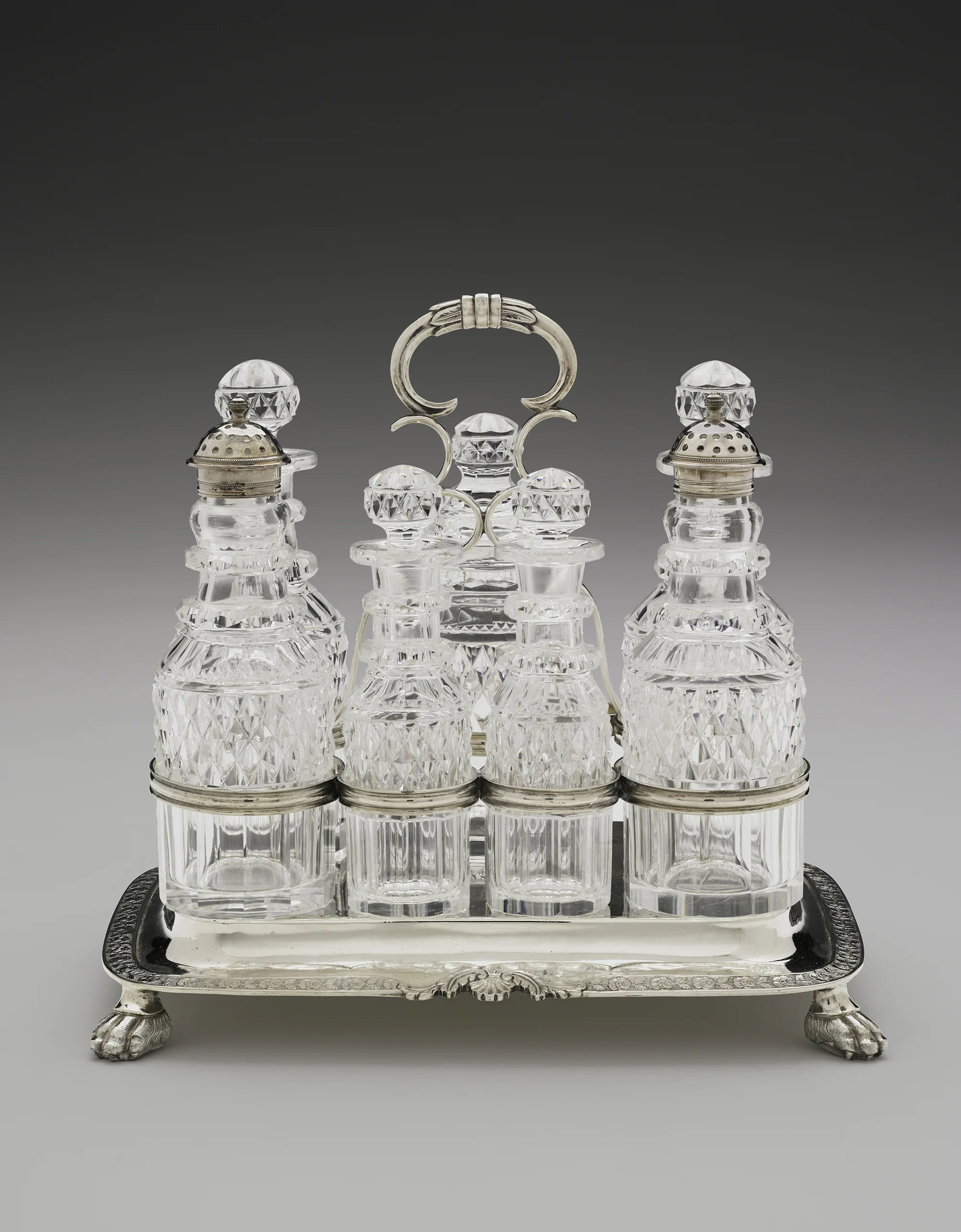 Elegant silver and glass vanity set on a silver tray, consisting of perfume bottles with faceted glass stoppers, and a silver jewelry holder with a decorative handle, all positioned on a white surface with a gray gradient background.