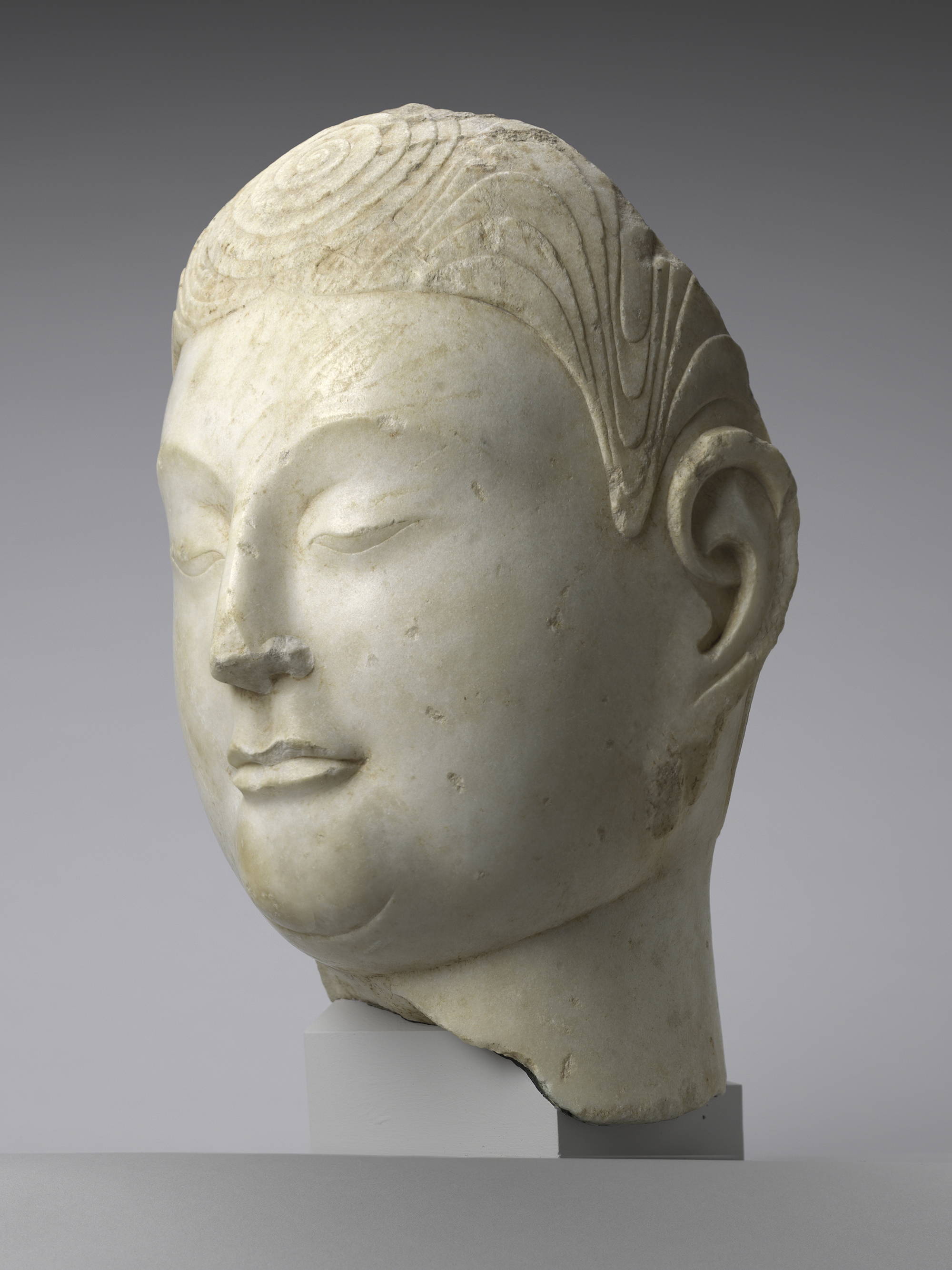 Ancient marble sculpture of a serene Buddha head with detailed carved hair and closed eyes, mounted on a display stand against a plain background.