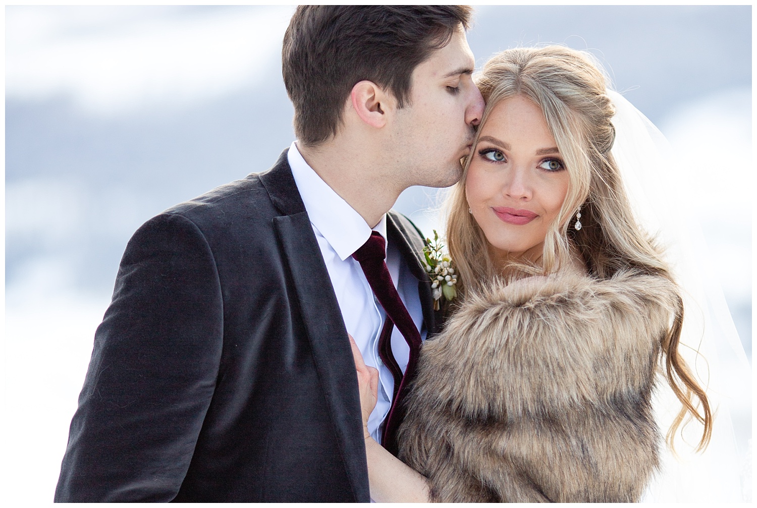 A groom kisses his bride on the temple while she smiles and looks away