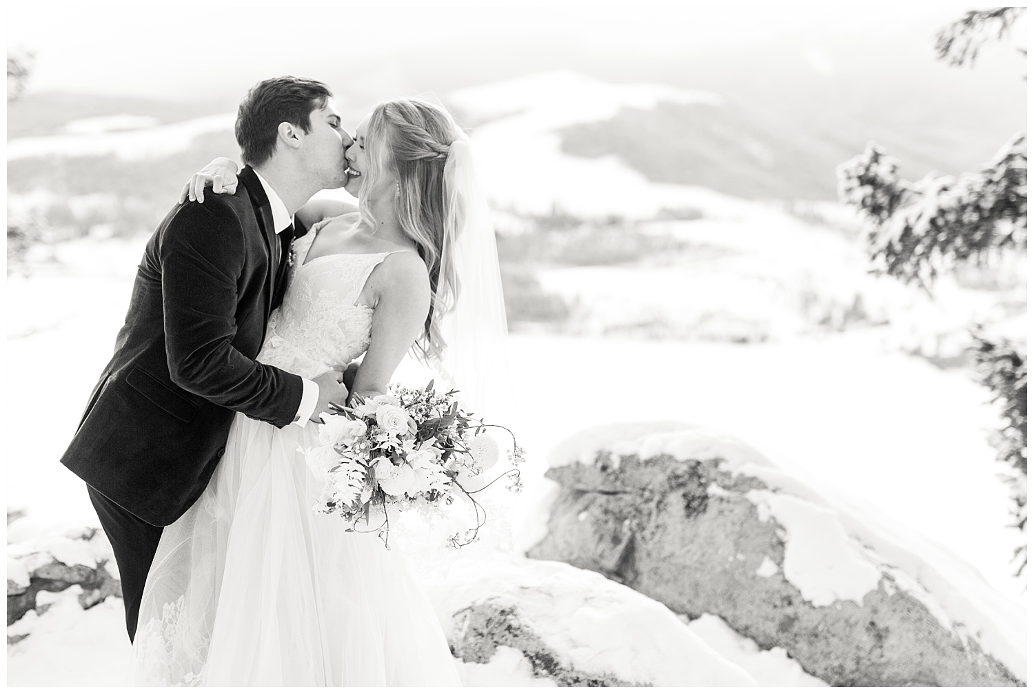 A groom helps his bride off of a rock and kisses her while she smiles