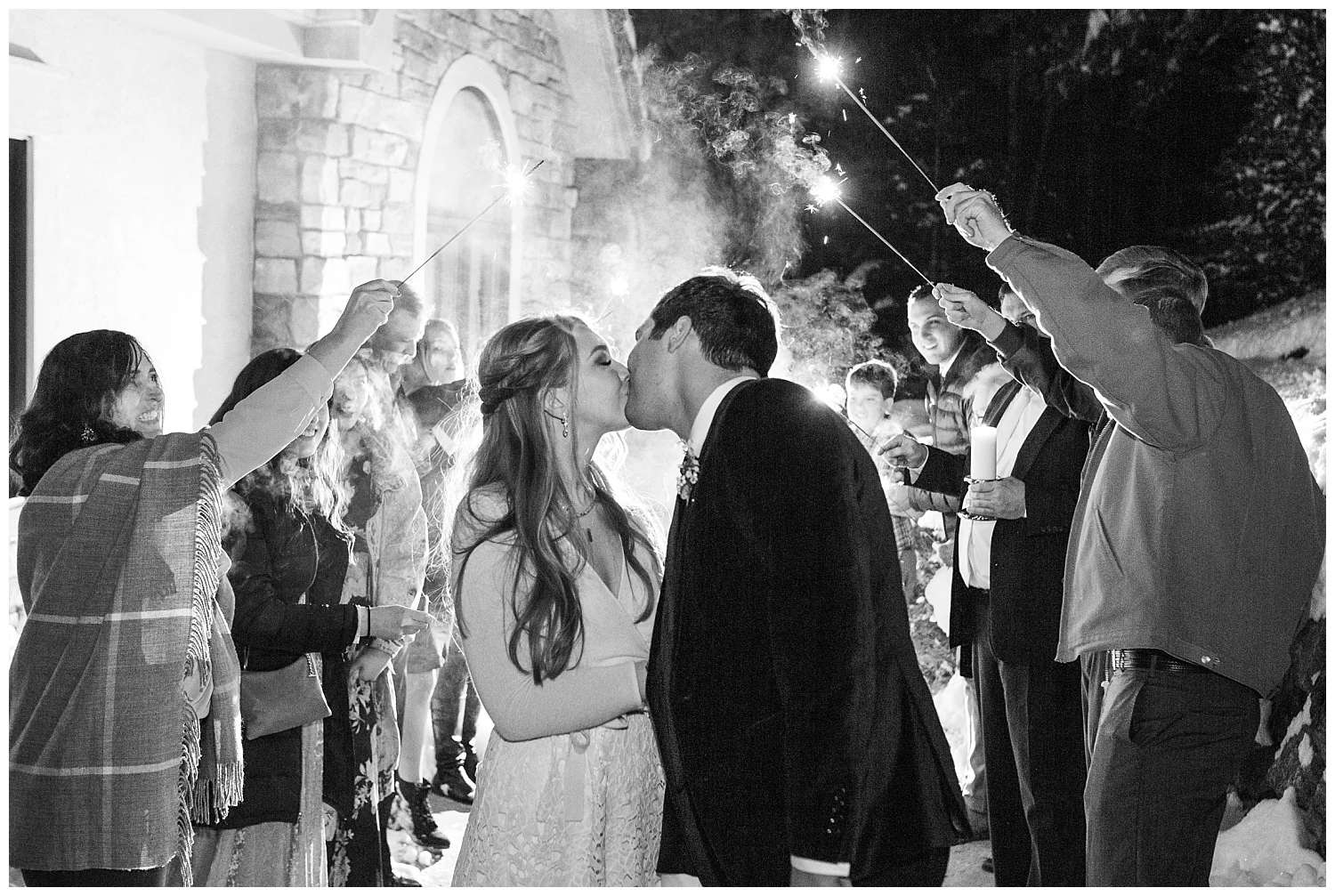 The bride and groom share their last kiss of the evening while the guests all hold sparklers