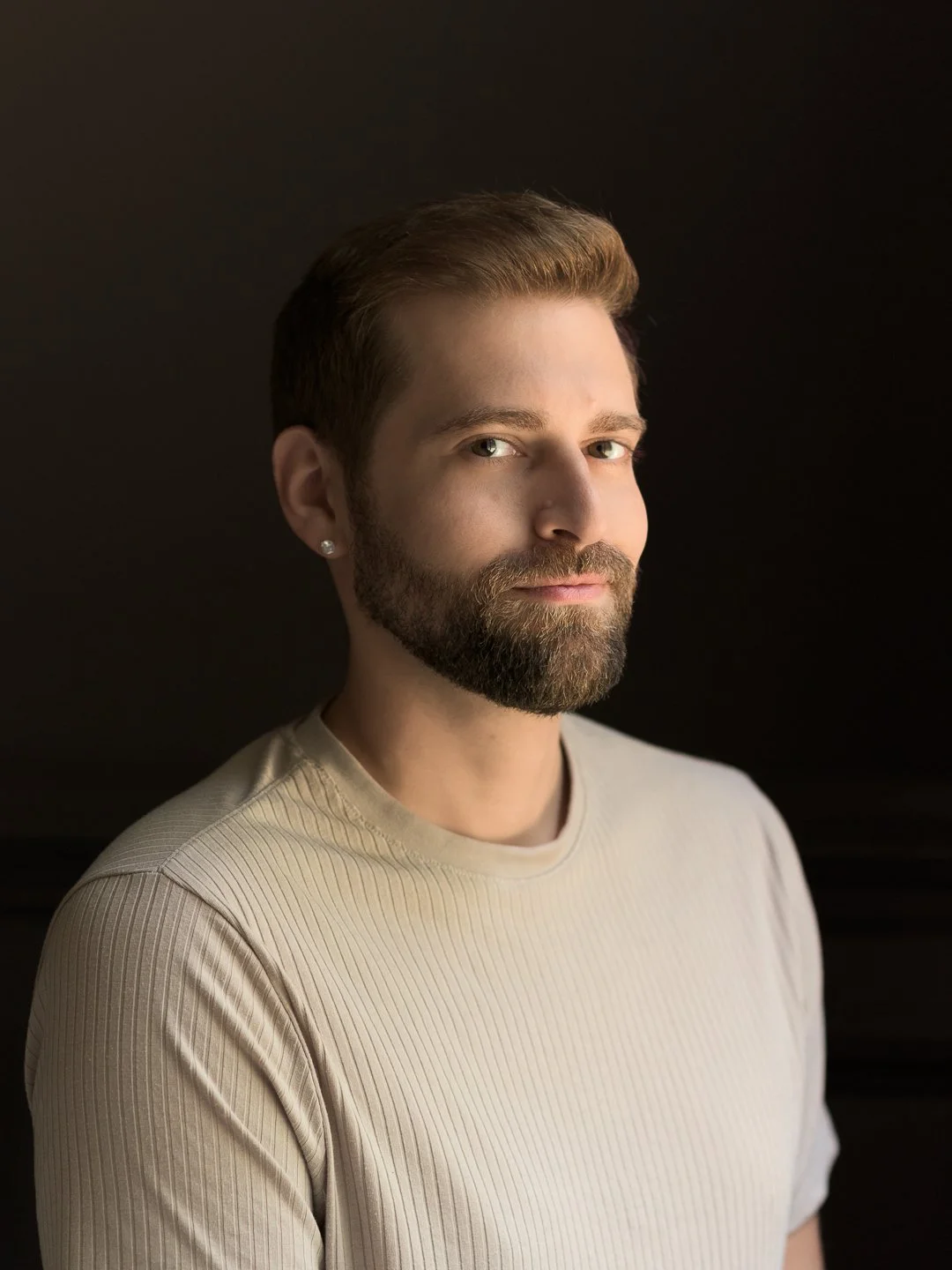 A young man with a well-groomed beard and styled hair, wearing a beige ribbed shirt, poses against a dark background, looking into the camera with a neutral expression.