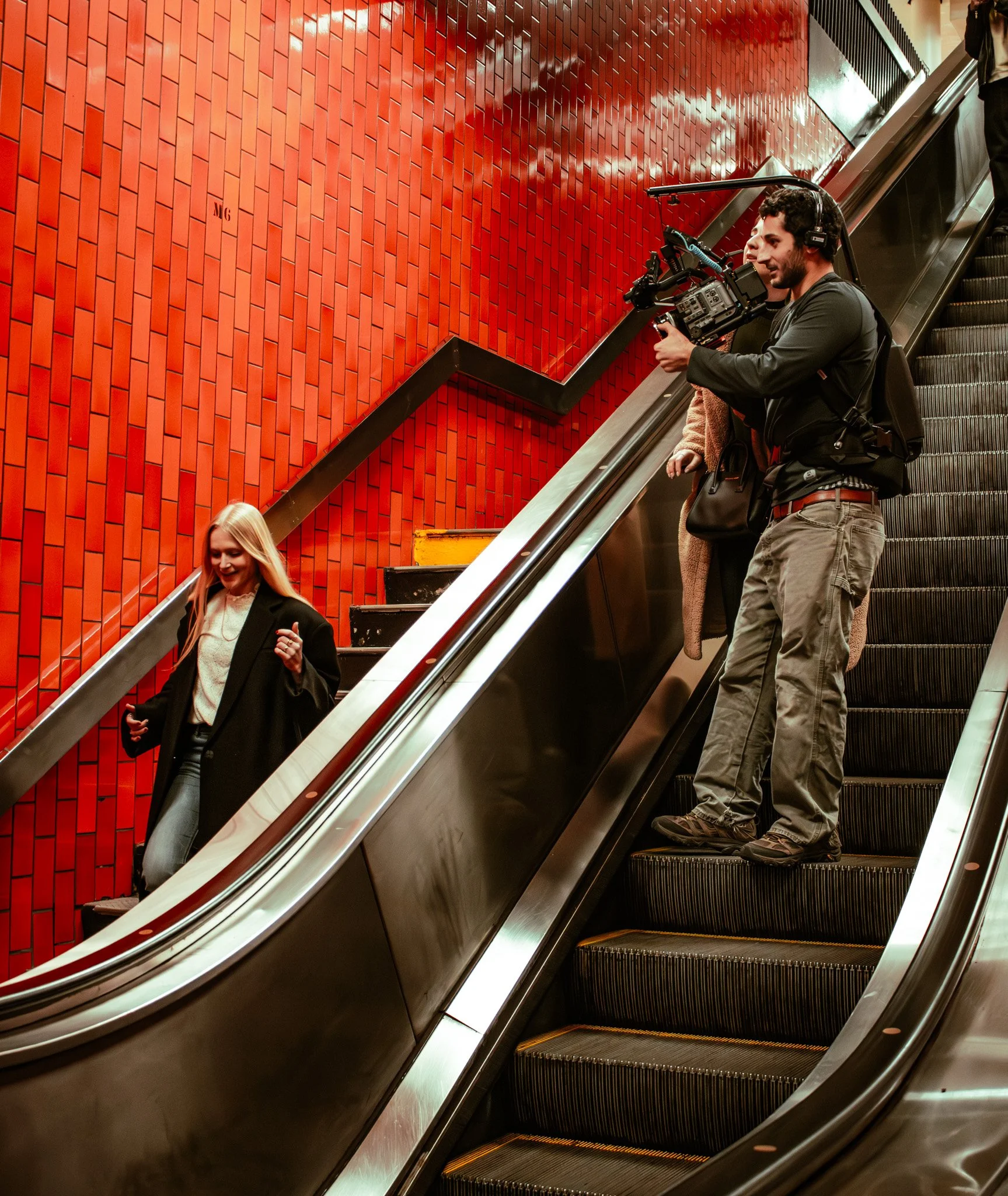 BTS of Drew Soleiman filming a woman descending an escalator in a subway station with red tiled walls.