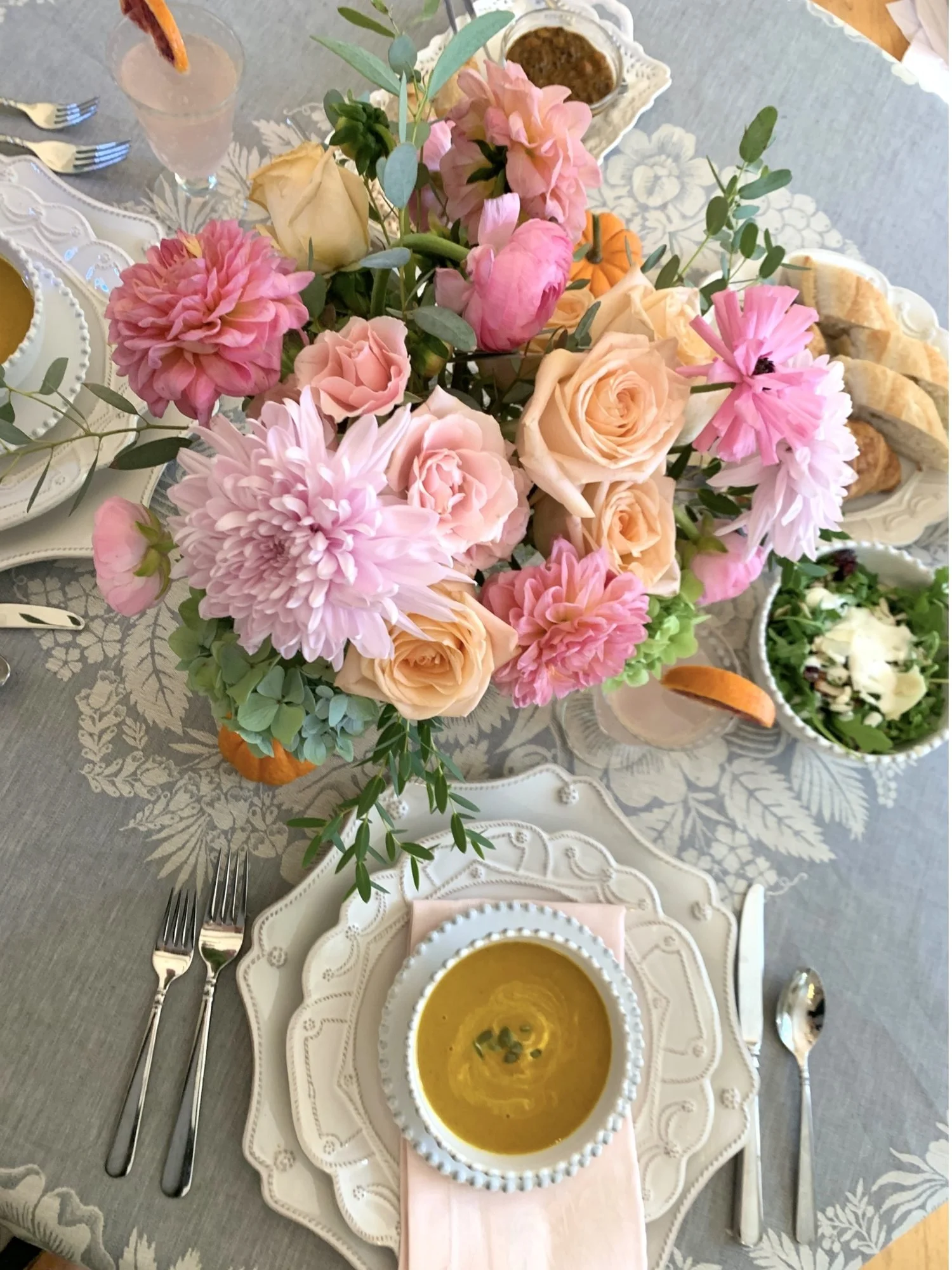 Overhead view of an autumn table with pink dahlias, peach roses and hydrangeas, golden soup in a white beaded bowl, and tiny pumpkins