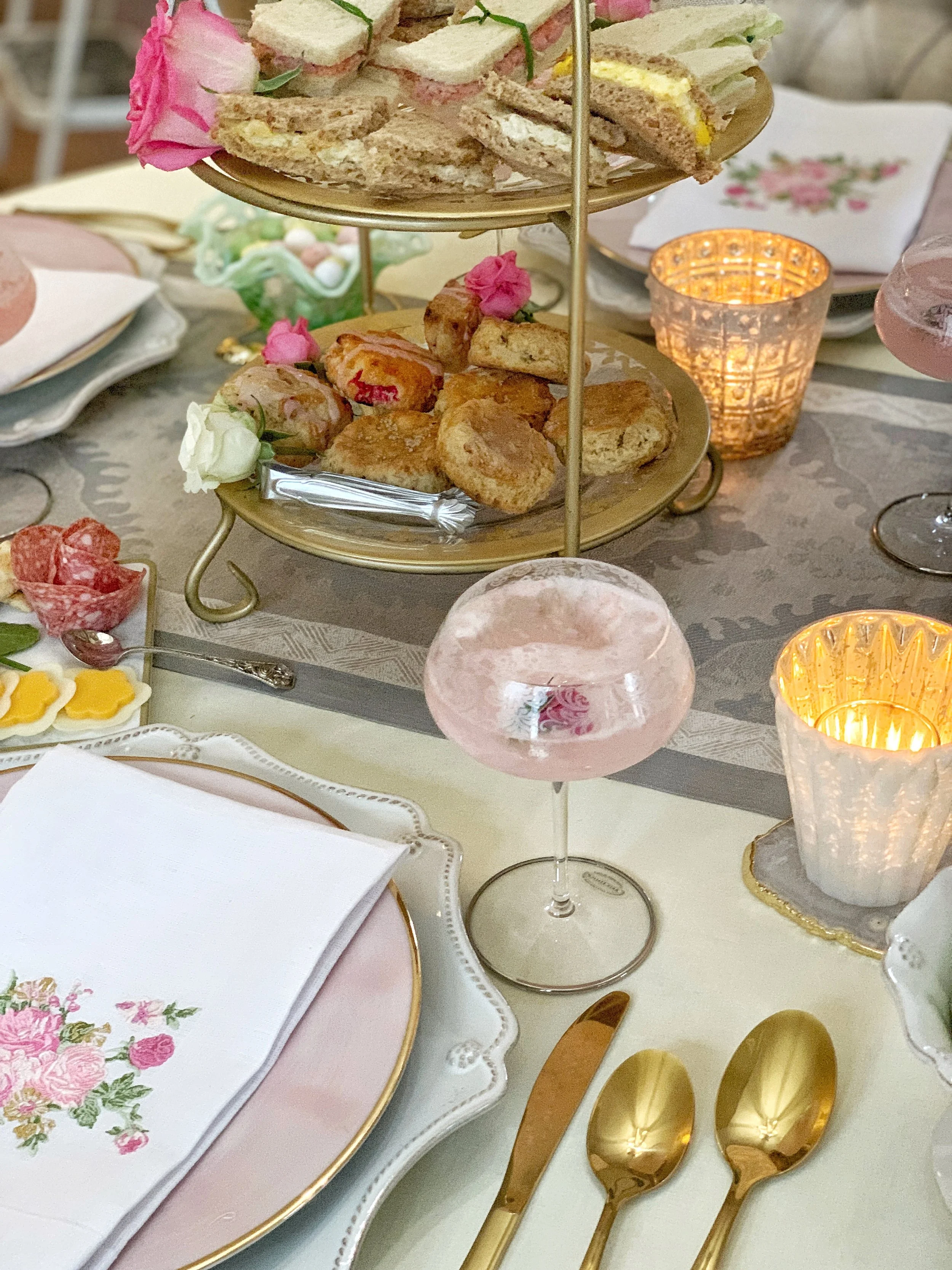 Elegant Easter high tea table with tiered serving stand, floral centerpiece, mixed china place setting, and glowing votive candles on a soft grey tablecloth.