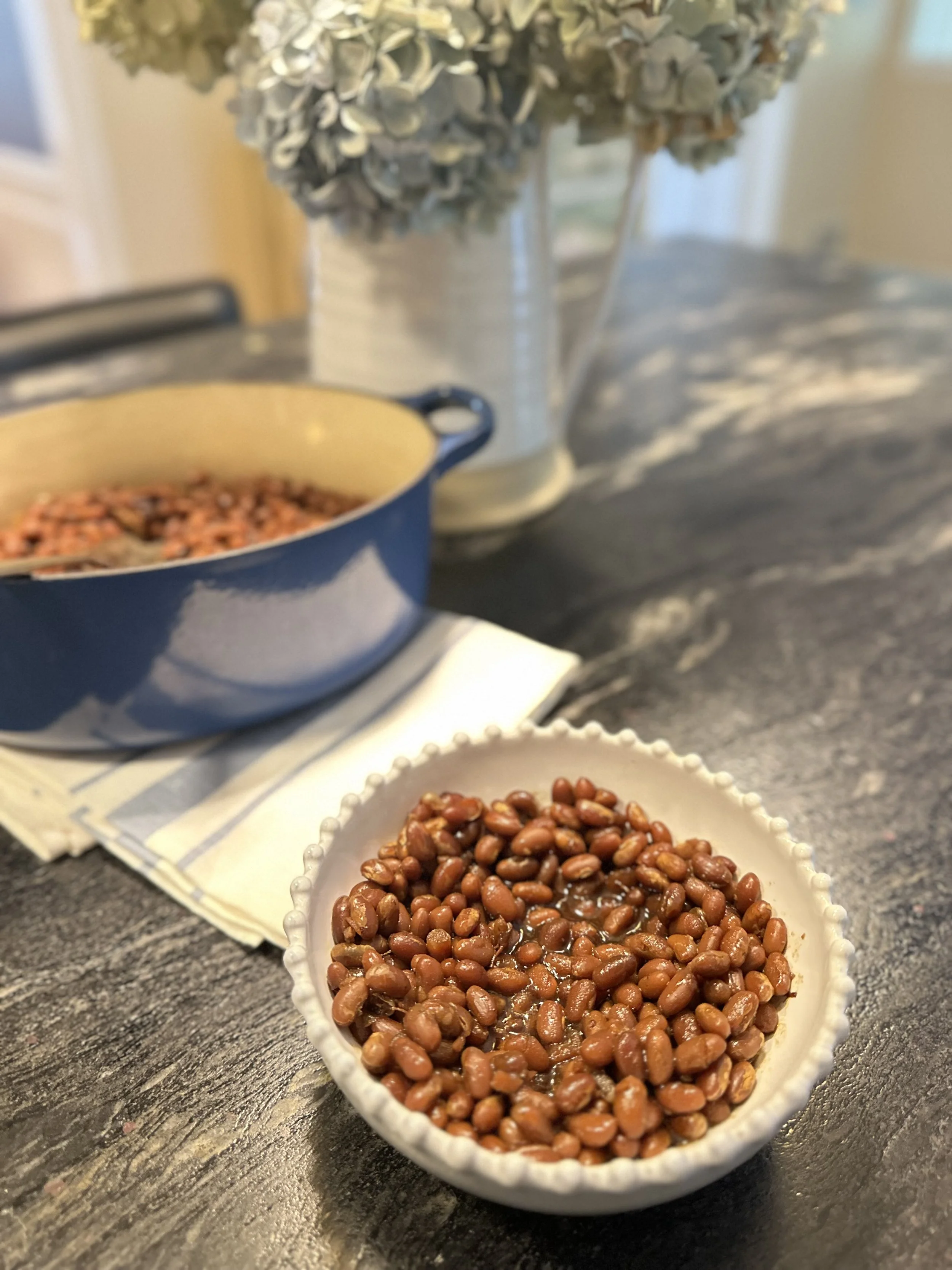 A white beaded bowl of homemade New England baked beans with a blue Le Creuset pot and hydrangeas behind