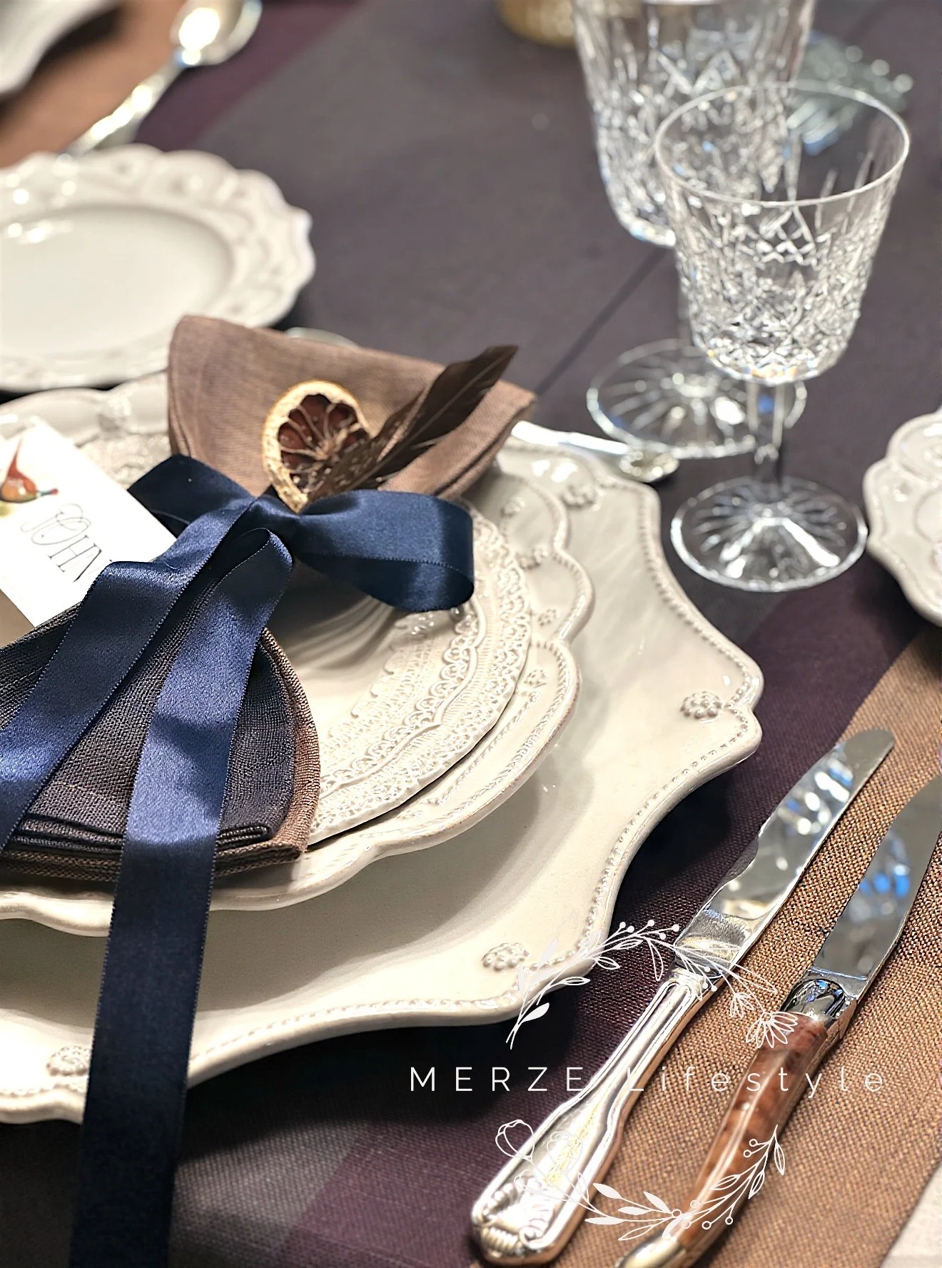 Cream table setting layered with patterned plates, woven linen napkin, and satin blue ribbon, styled with sterling silver flatware and crystal glassware for a quiet luxury tablescape.