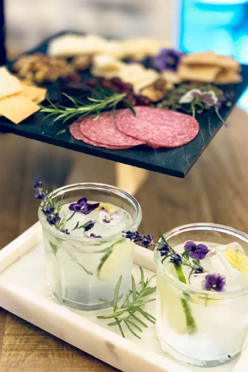 St Germain lavender gin and tonic garnished with edible flowers served beside a summer cheese board with fruit, nuts, and honey.