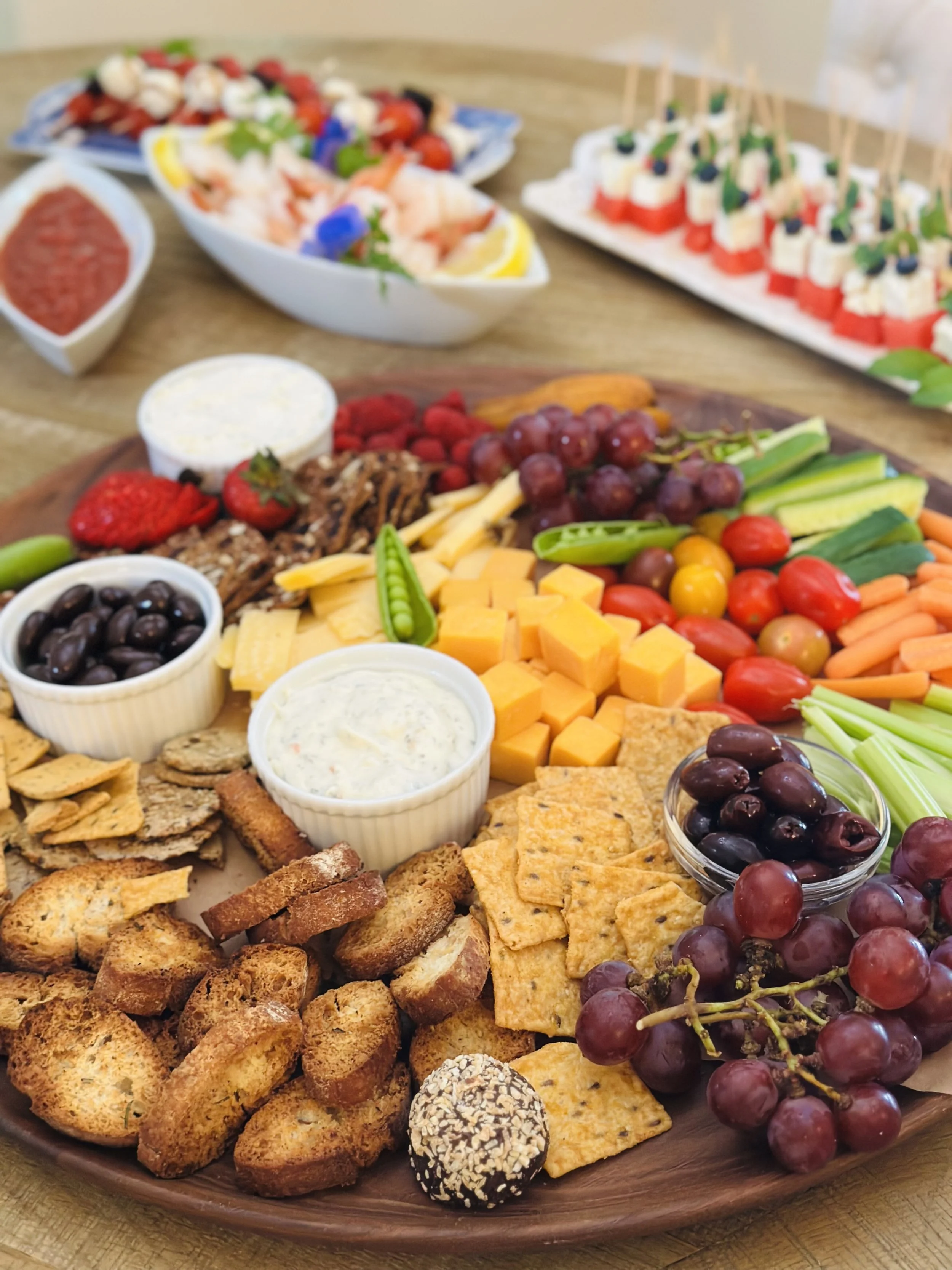 Summer charcuterie grazing board with cheeses, fresh fruit, raw vegetables, nuts, crackers, olives, and herbed dips on a round wood board.