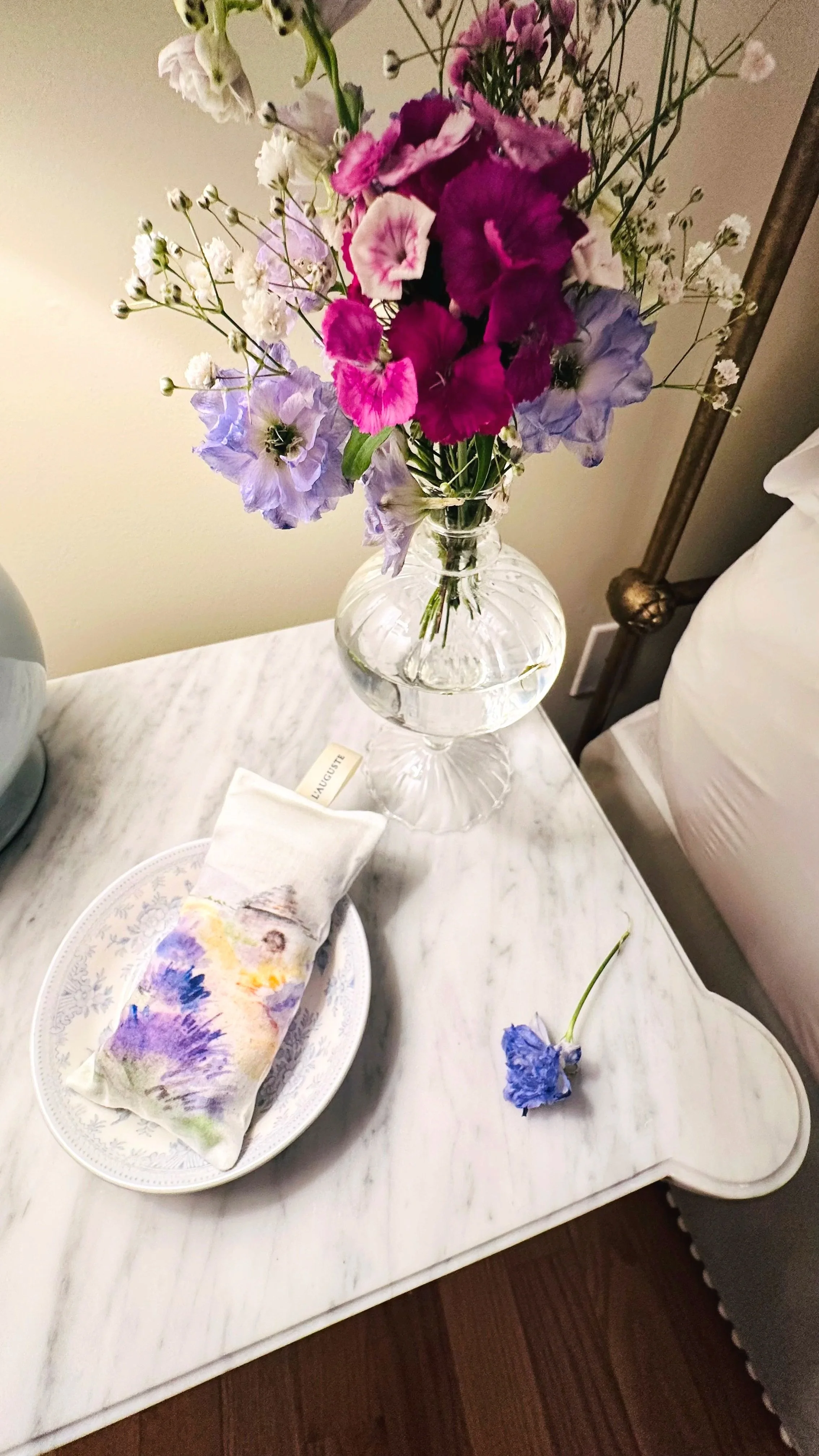 A marble-topped nightstand with a bud vase of spring wildflowers, a blue and white dish holding a lavender sachet, and a floral lamp.