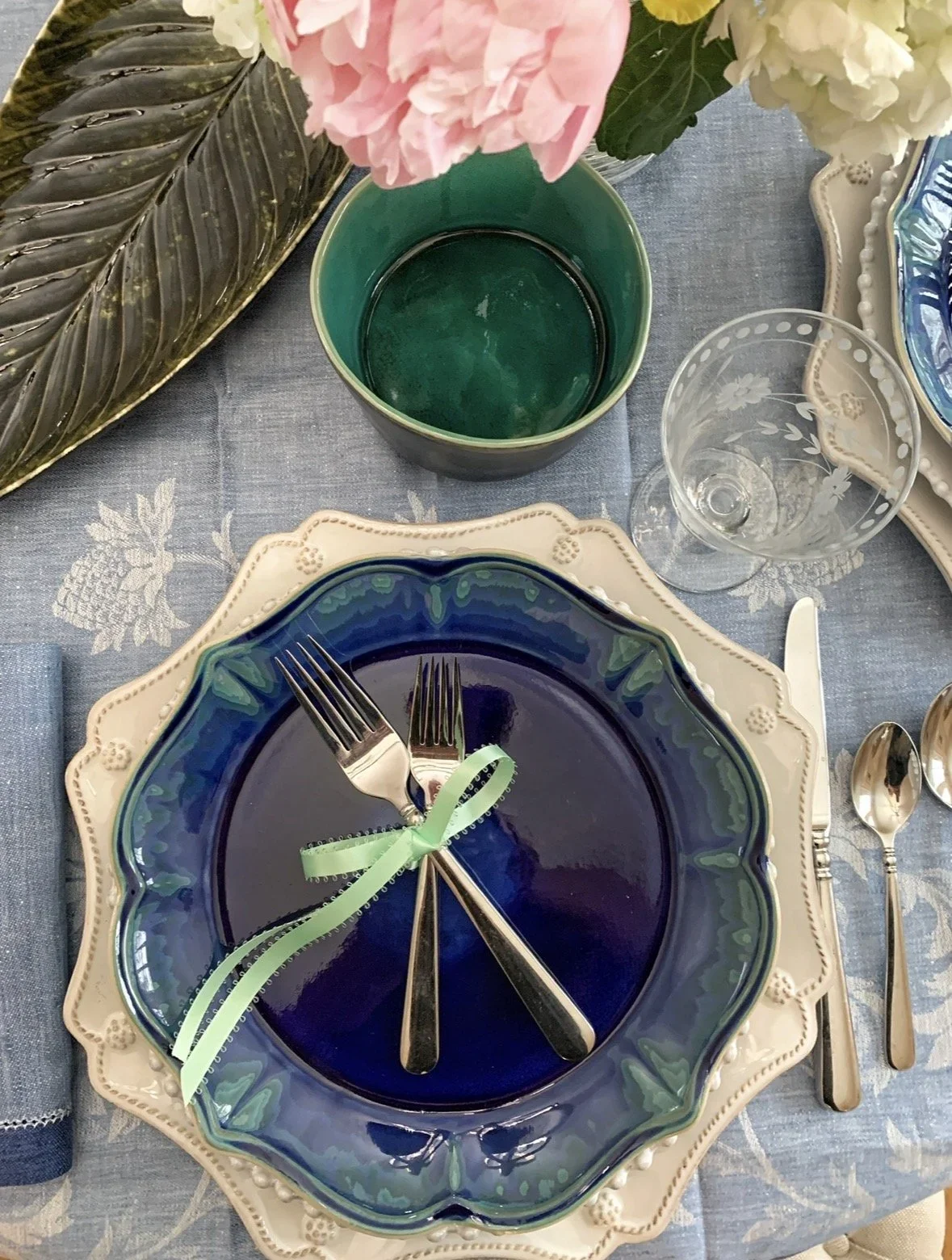 Coastal jewel-tone place setting with a cobalt and teal glazed plate, scalloped charger, and green ribbon detail