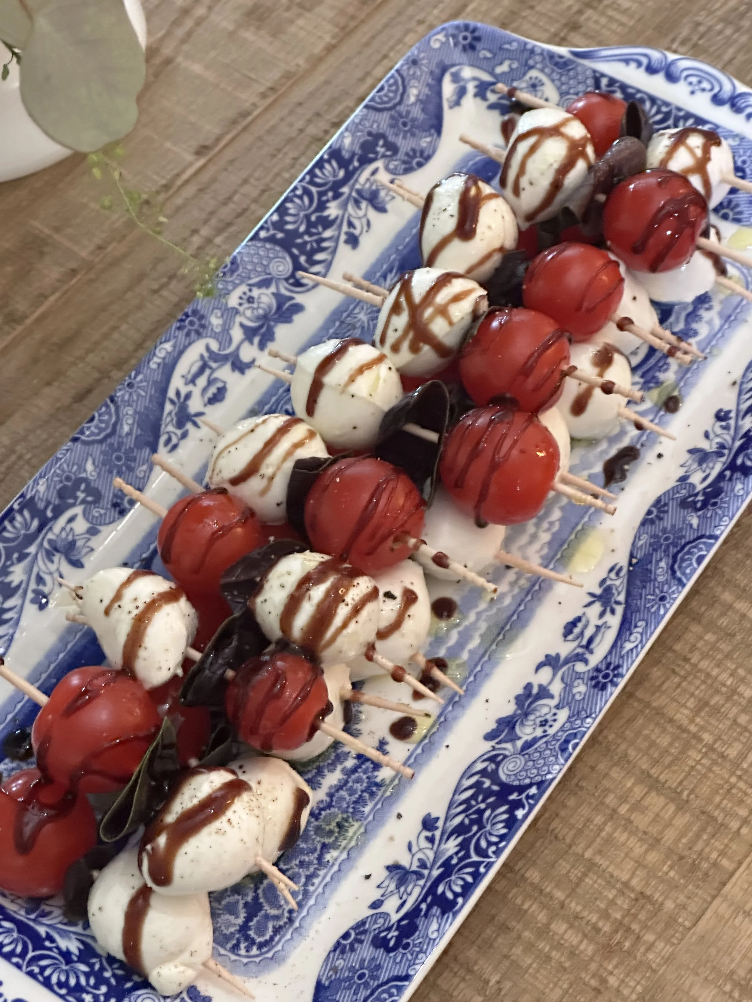 Caprese skewers with cherry tomatoes, fresh mozzarella, basil, and balsamic glaze on a blue and white transferware platter.