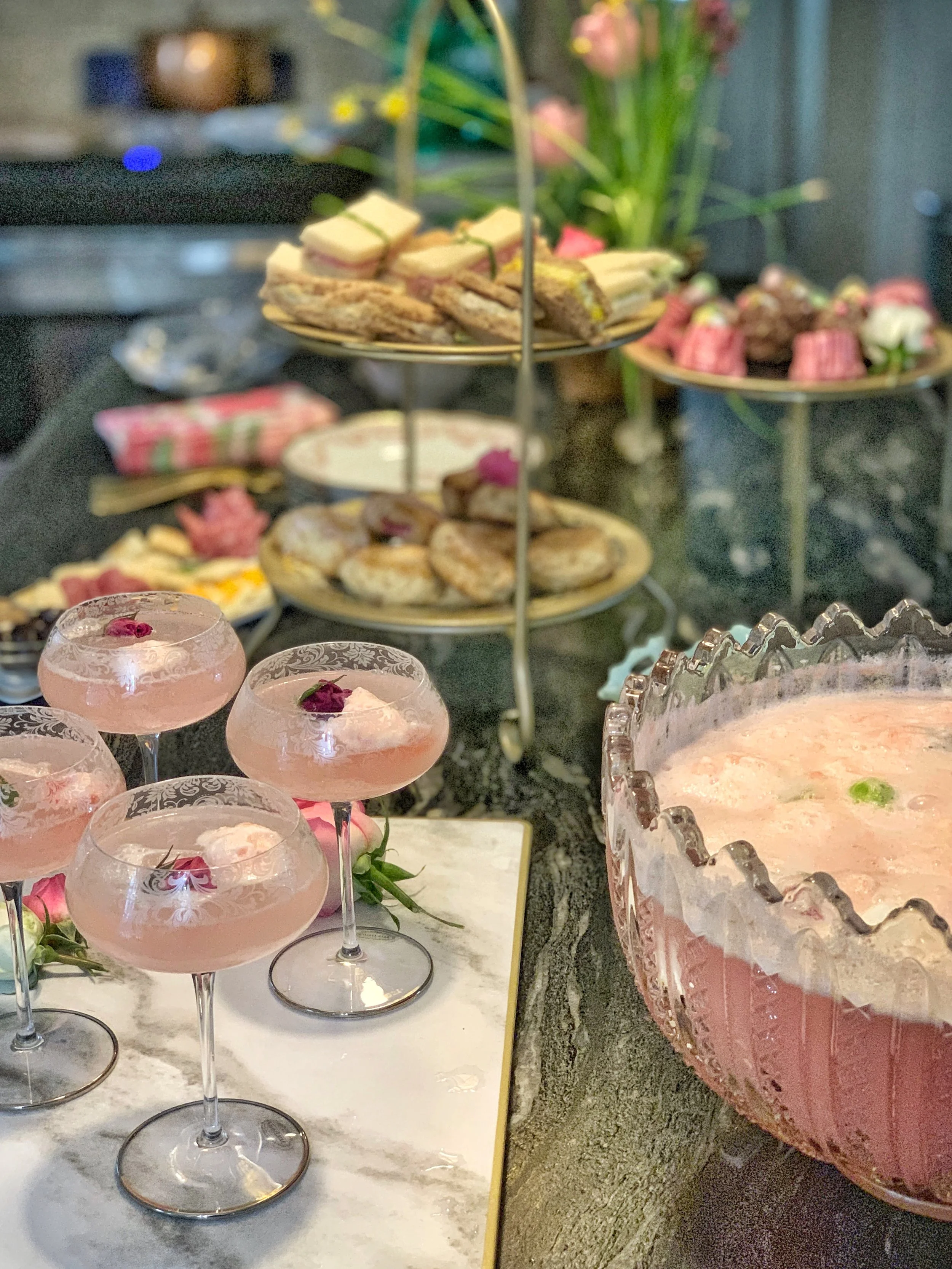 Easter high tea display on a kitchen counter featuring tiered stand with desserts, vintage punch bowl, and etched wine glasses filled with a rose-infused spring drink.