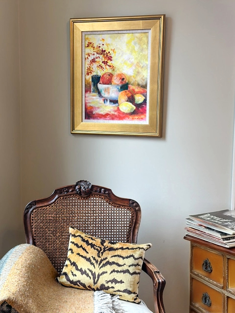 Studio vignette with antique French chair, linen blanket, Scalamandre pillow, magazines, and a painting created by the author's mother, representing creative inspiration and returning to art later in life.