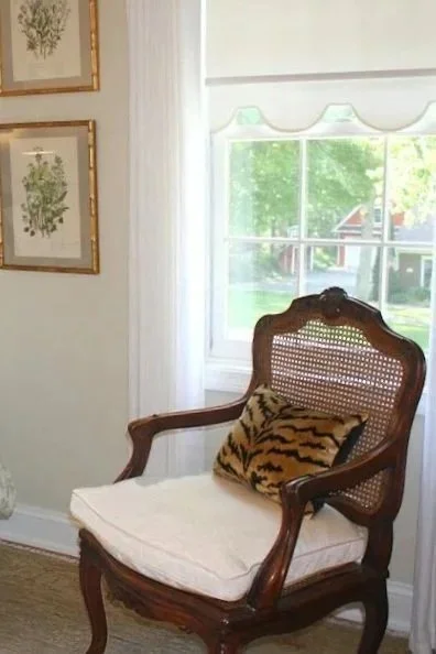 An antique French cane-back chair with a tiger velvet pillow beside a sunlit window, with framed botanical prints on the wall above.
