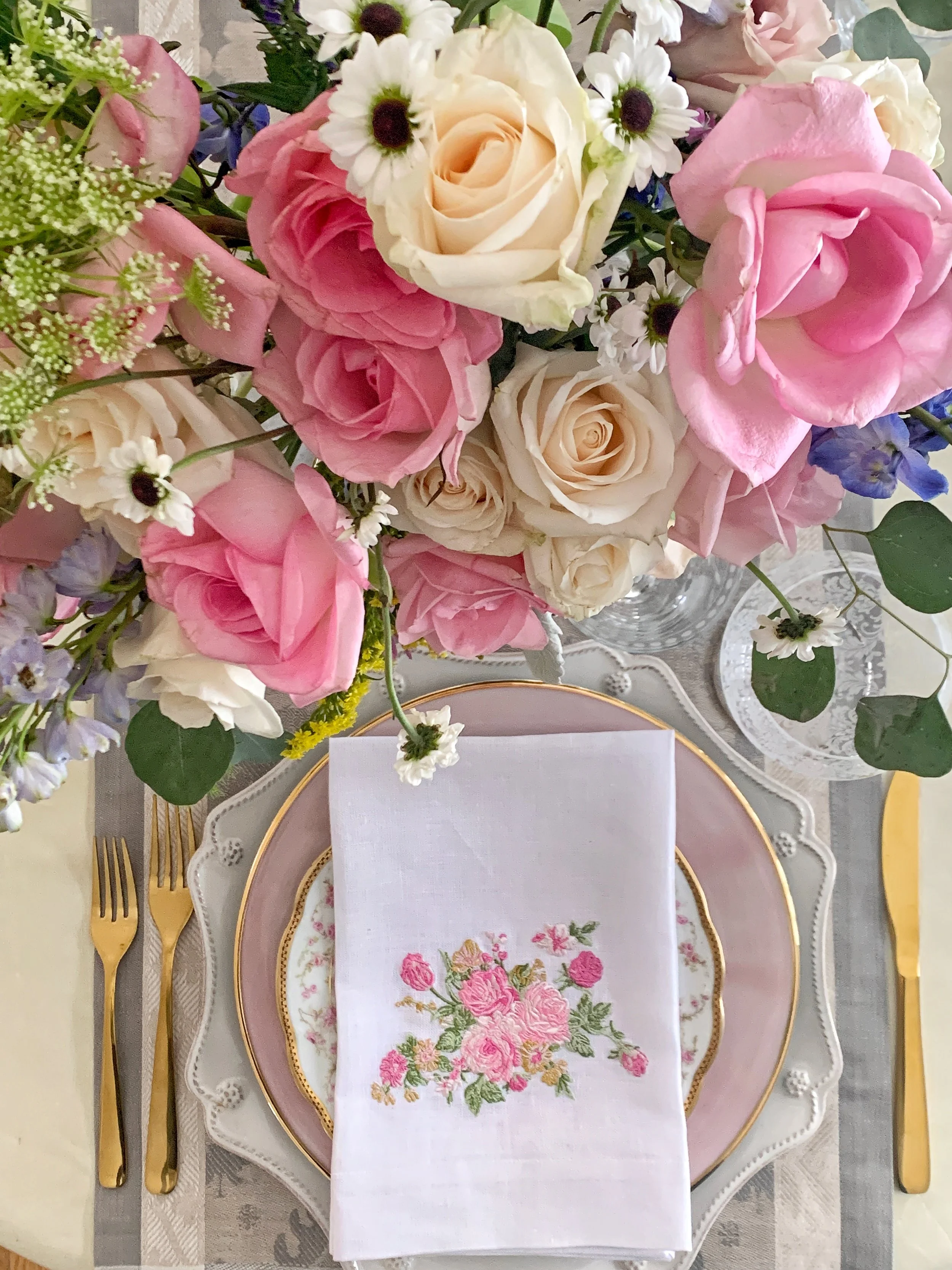 Spring Easter high tea table featuring layered vintage and modern china, embroidered floral linen napkin, gold flatware, etched glassware, and fresh seasonal flowers.